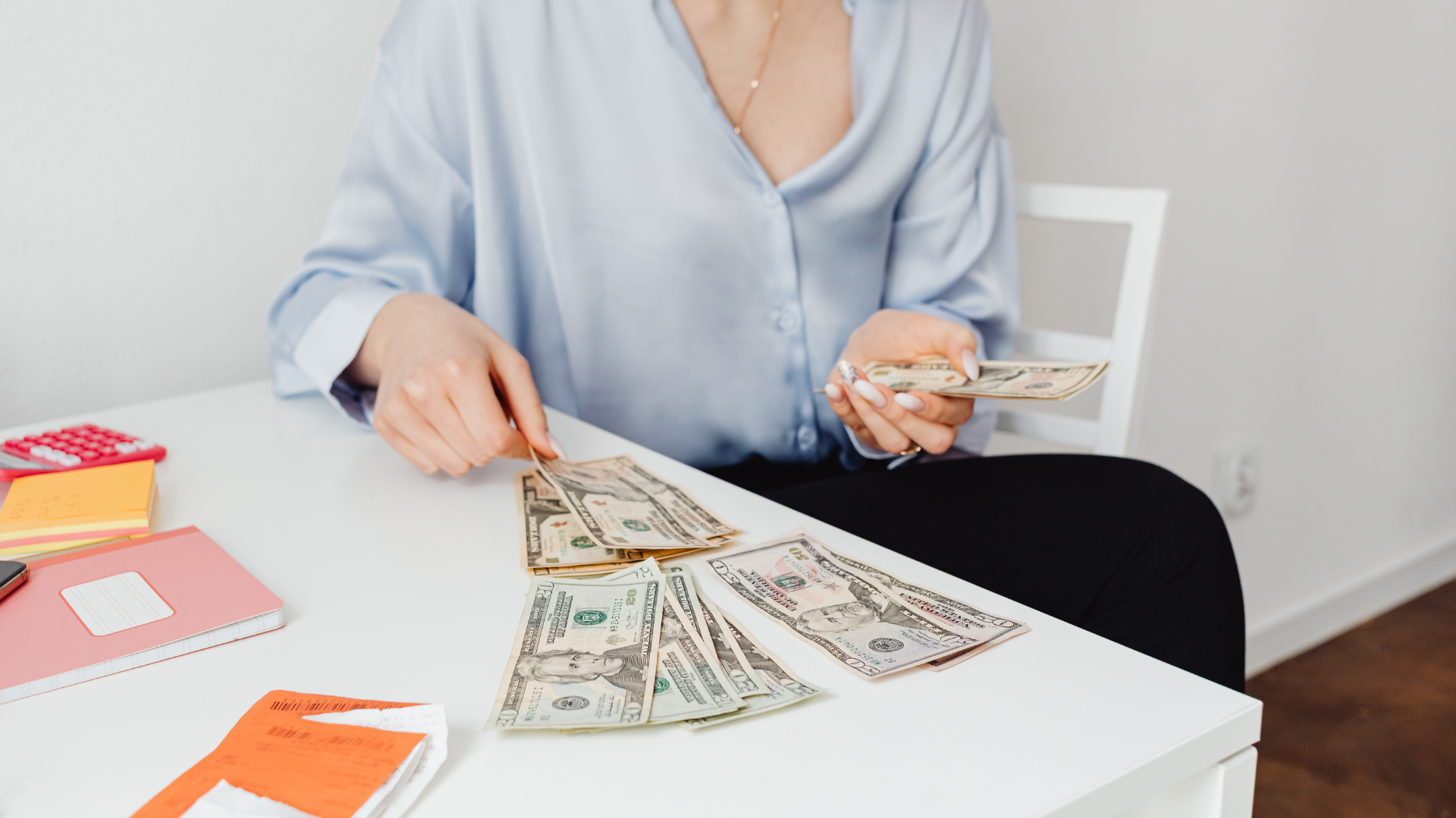 Person counting dollar bills at a white table with paperwork and office supplies