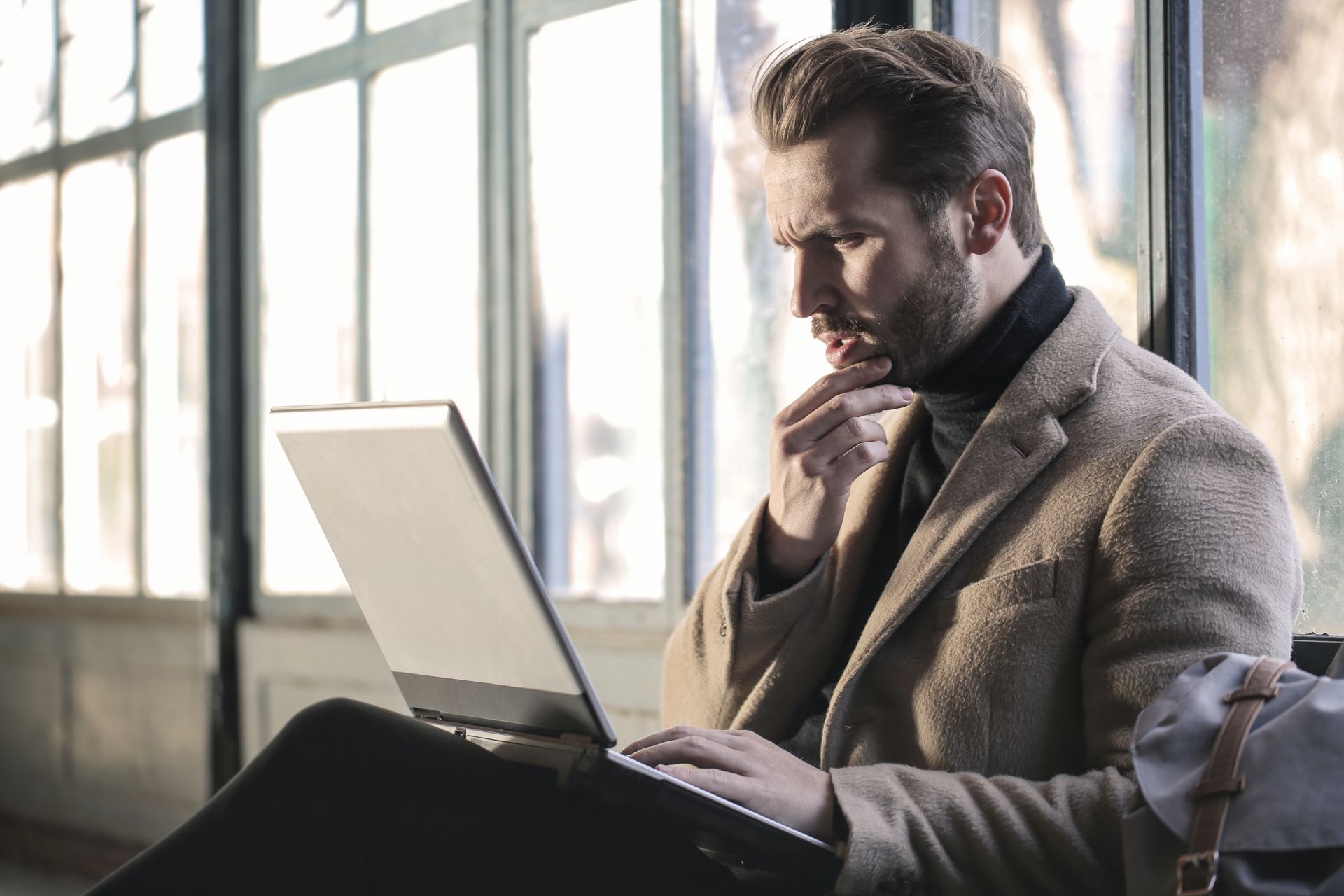 A man is sitting in front of a window using a laptop computer.