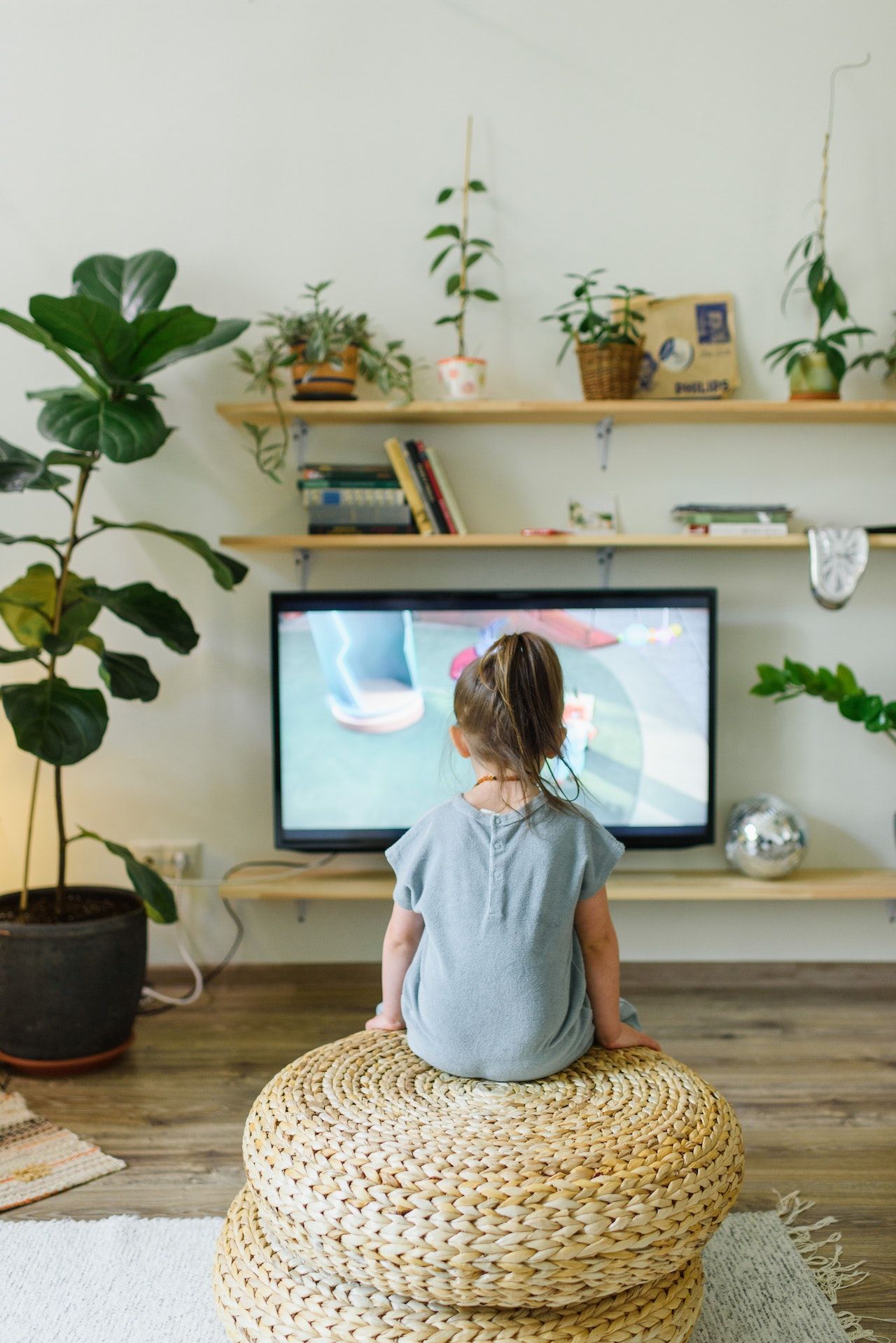 A little girl is sitting on a wicker ottoman in front of a television.