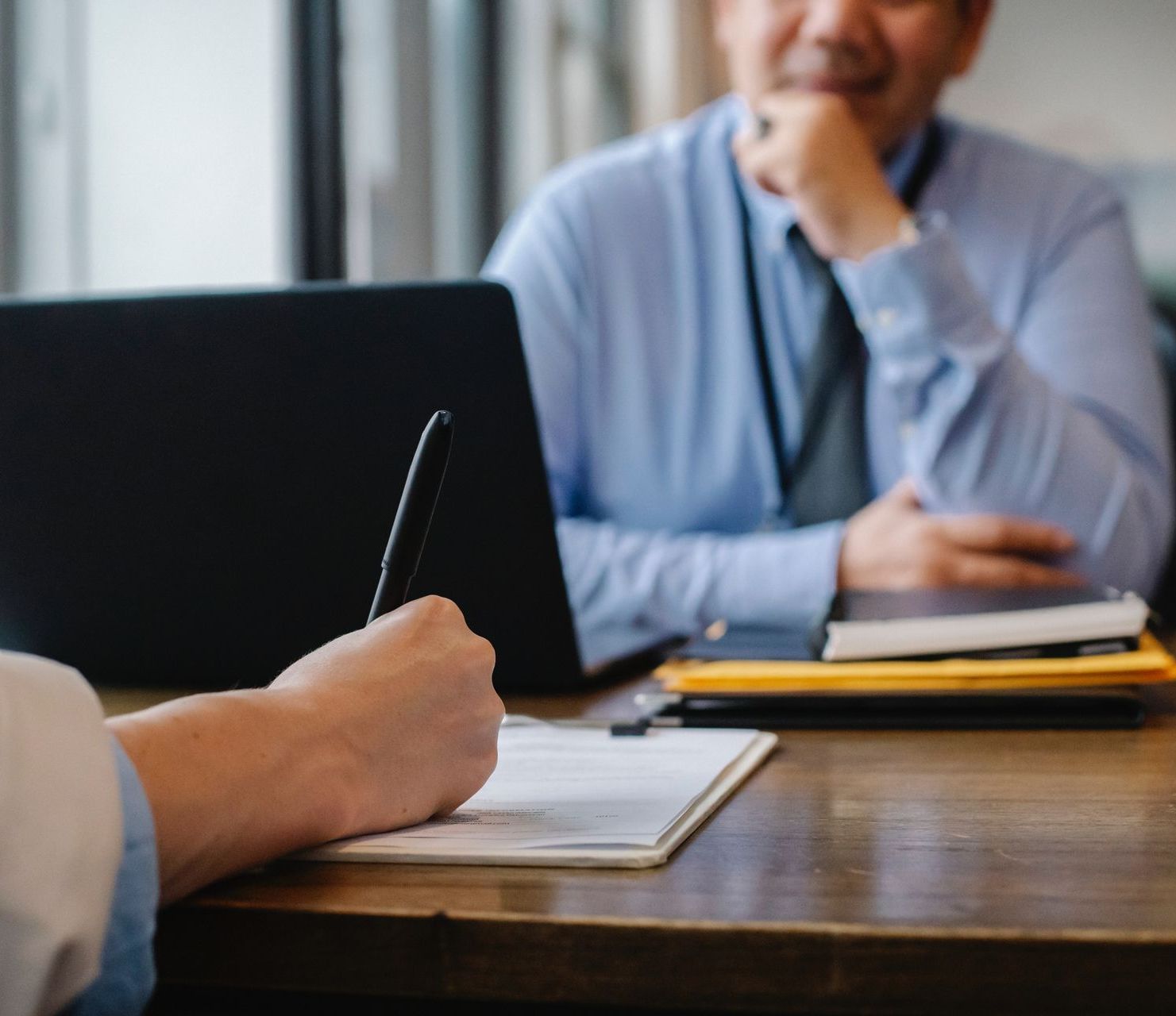 A man is sitting at a desk with a laptop and a woman is writing on a piece of paper.