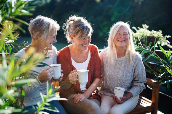Three older women are sitting on a bench holding cups of coffee.
