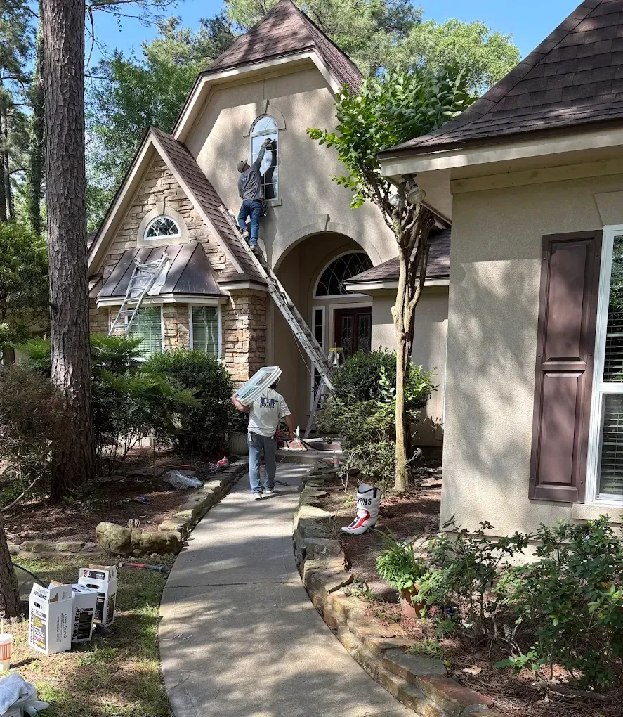 A worker on a ladder installs an upper window on a tan house with stone accents, while another person walks toward them.