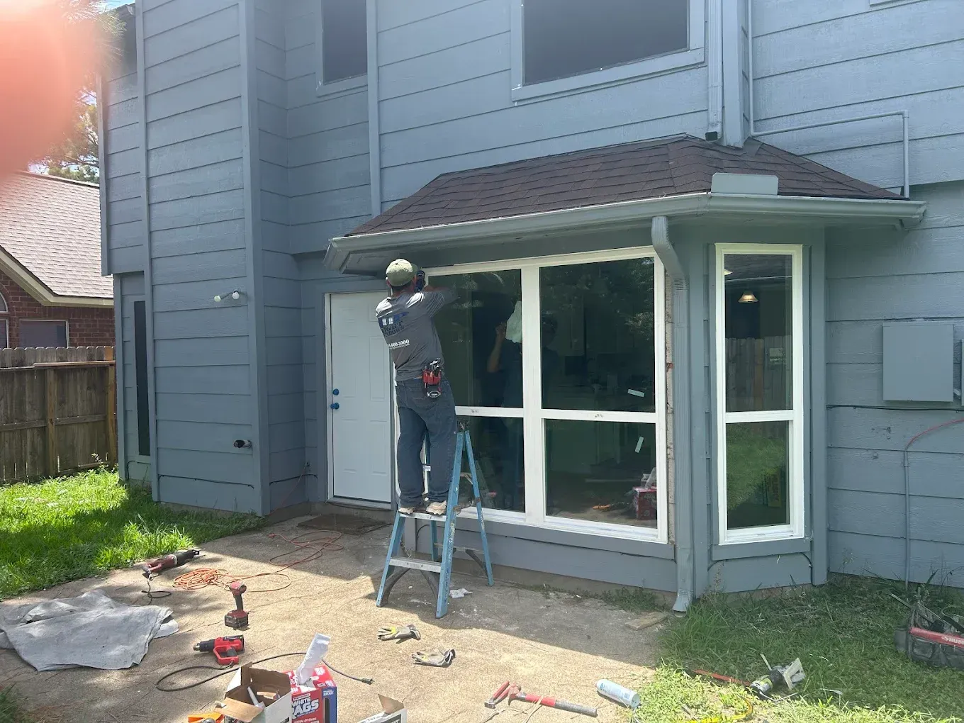 A person on a stepladder works on the windows of a grey, two-story house on a sunny day.