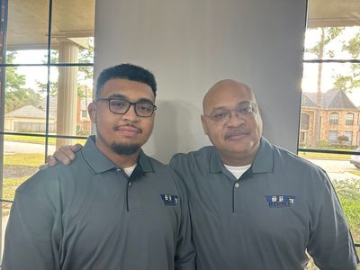 Two men in matching grey shirts stand side-by-side indoors near a window, smiling at the camera.