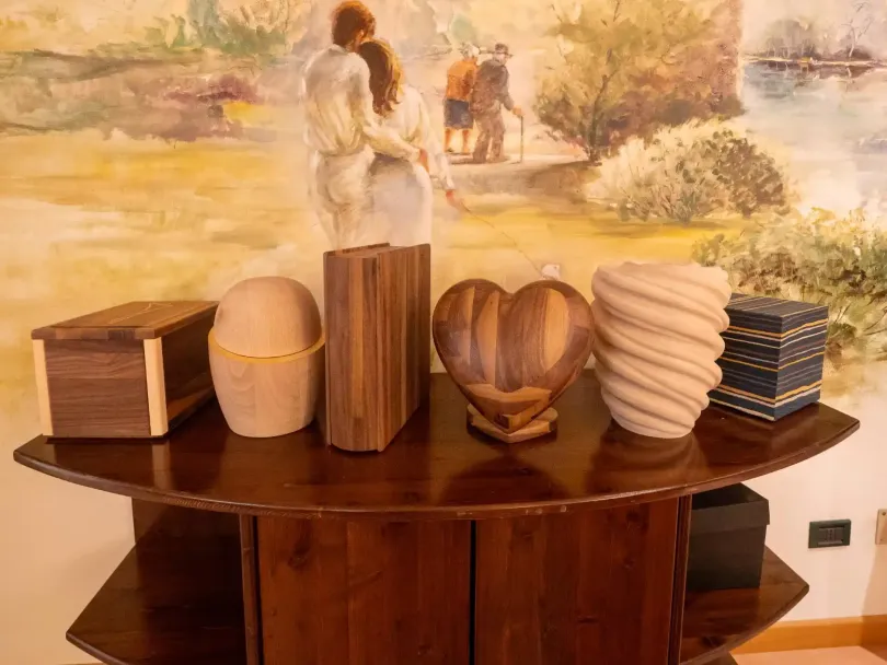 Urne di legno di varie forme e dimensioni su un tavolo di legno scuro, con uno sfondo murale raffigurante persone in un paesaggio.