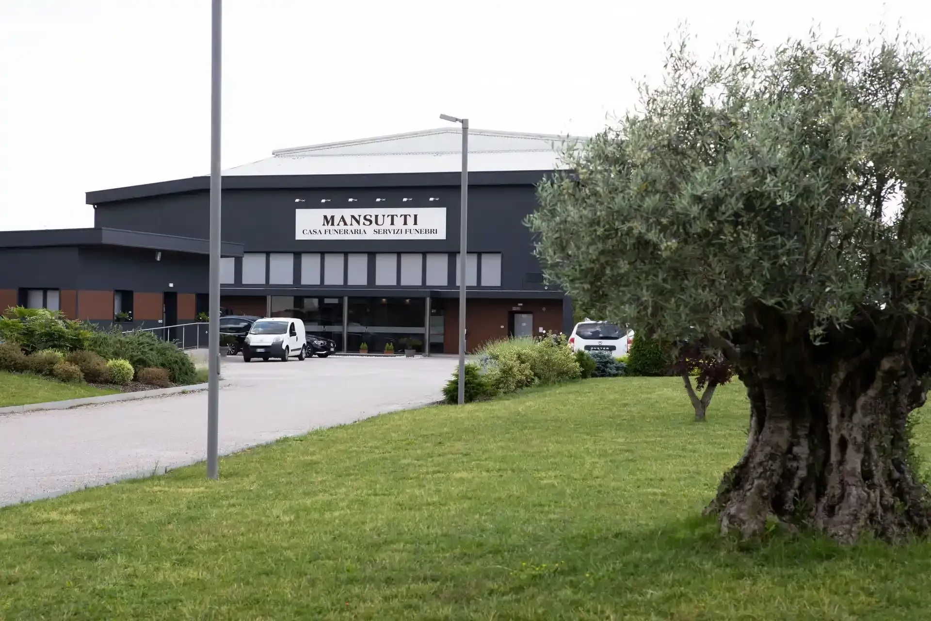 Edificio grigio scuro con un'insegna bianca, veicoli parcheggiati e un grande albero di fronte.