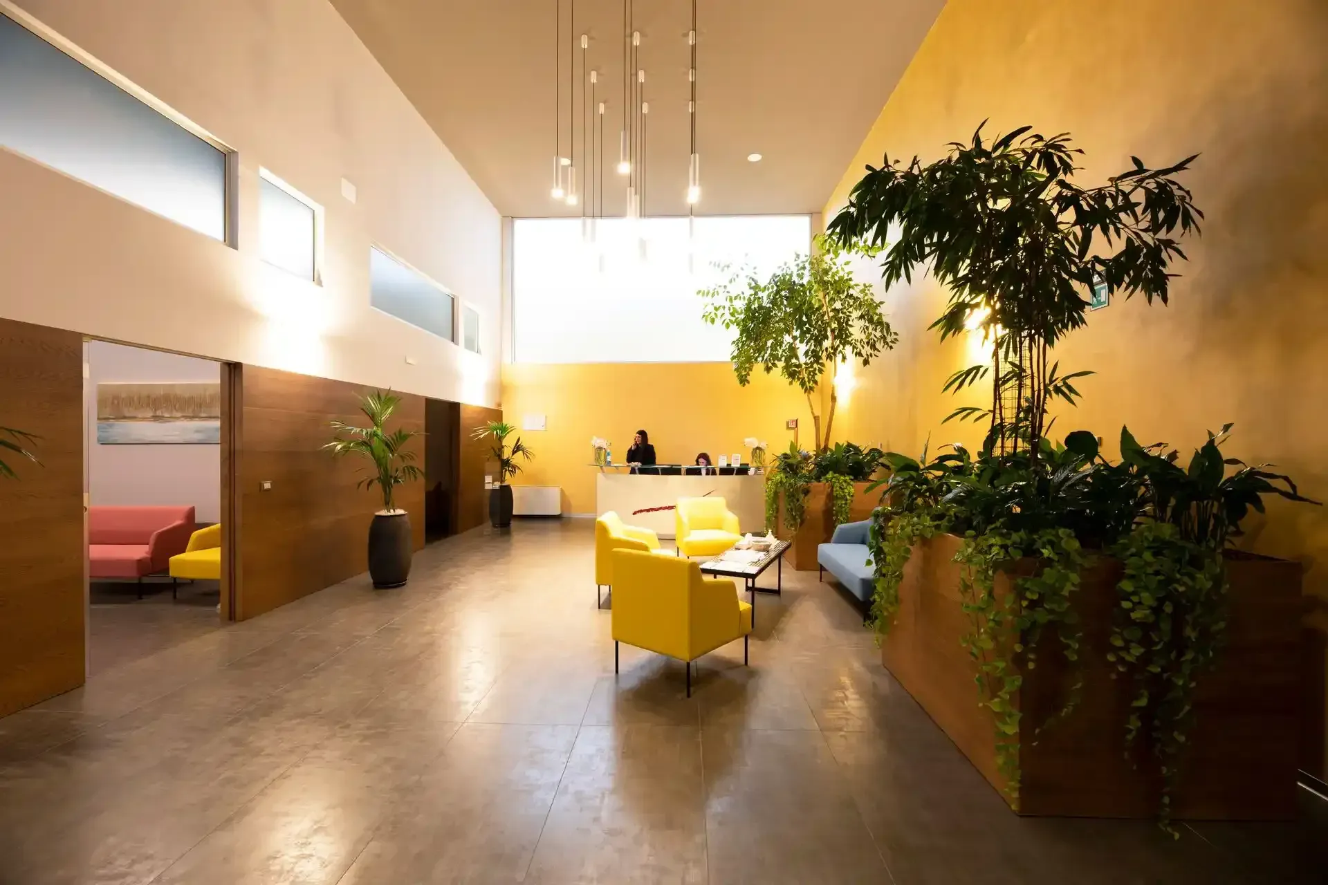 Hall luminosa con reception, poltrona gialla, piante e pavimento in cemento.