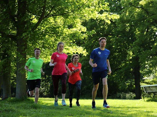Vier mensen rennen over een graspad in een park met bomen; ze dragen sportkleding.