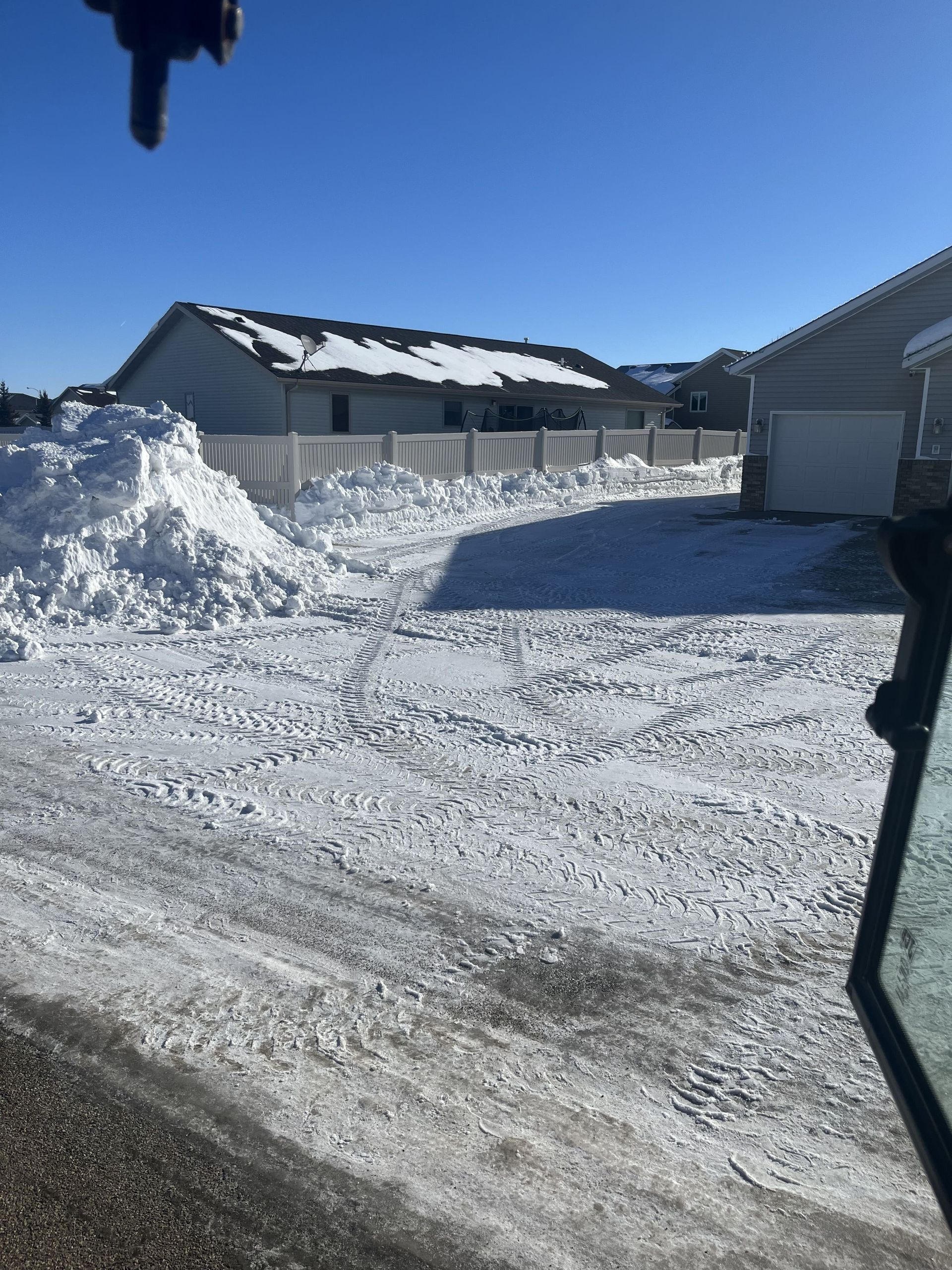 Snow-covered driveway with a large snow pile, clear sky, houses in the background.