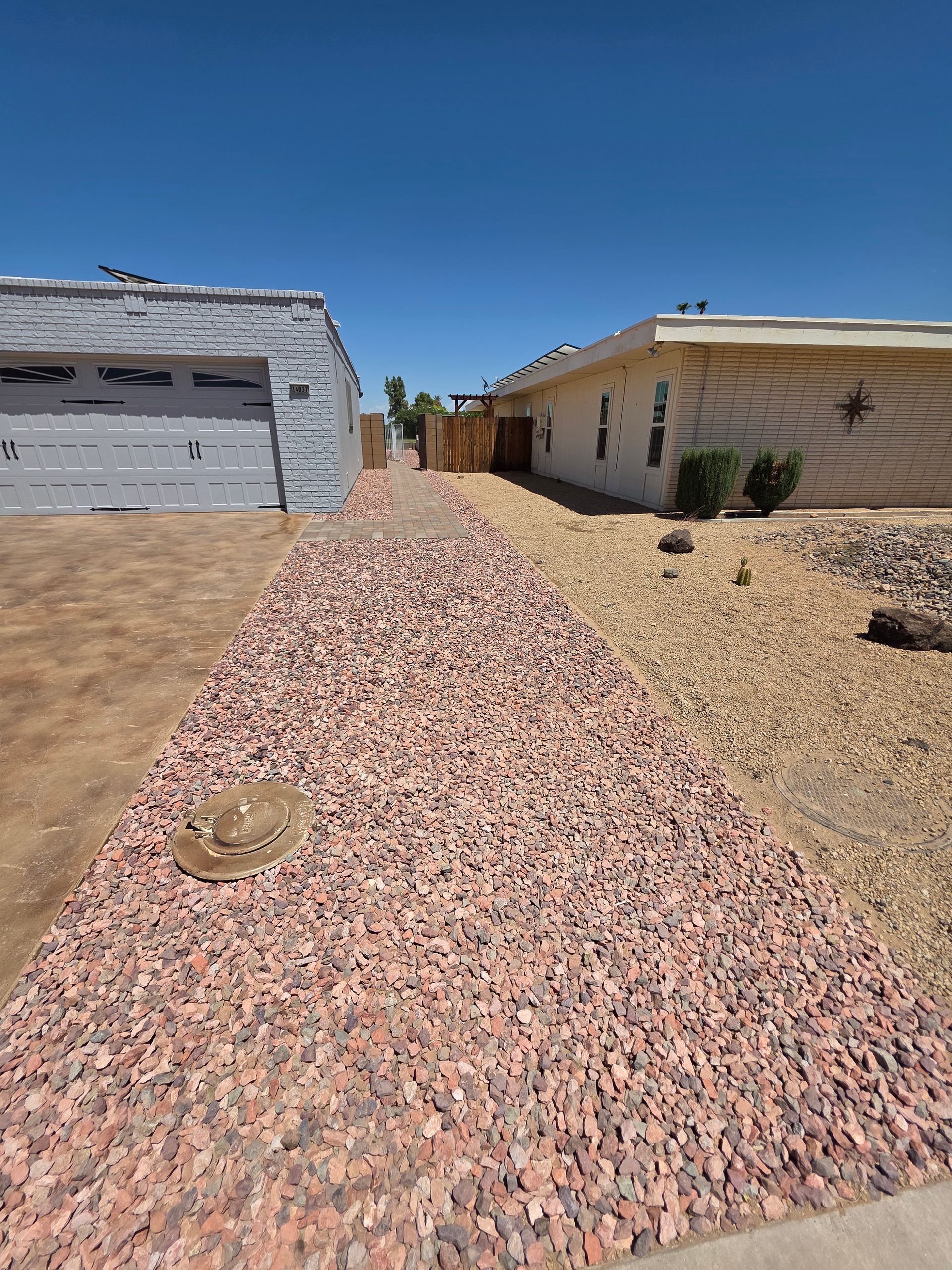 Path of reddish gravel between two houses under a clear blue sky. One house is off-white, the other has a grey garage door.