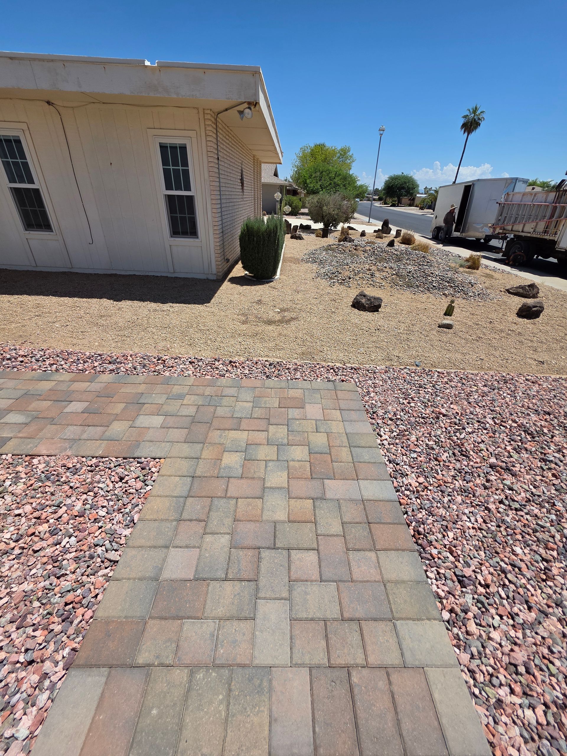 Brick walkway leading to a house with a gravel yard. A truck and palm tree are visible in the background on a sunny day.