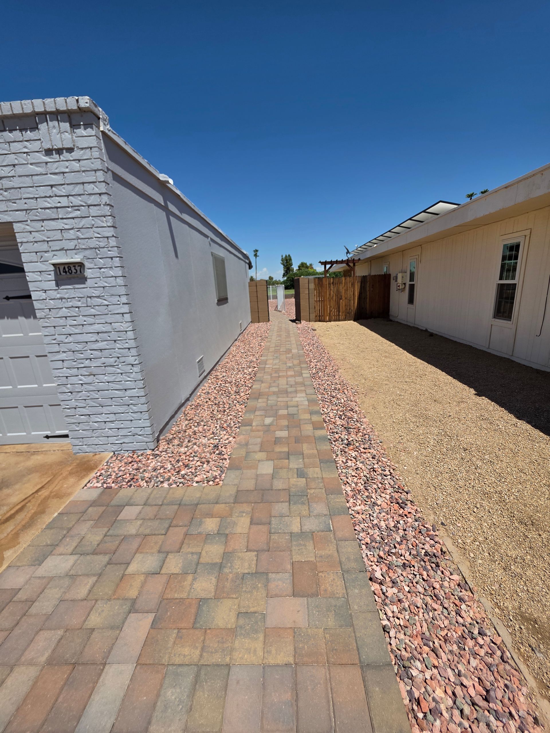 Narrow walkway between two buildings, paved with bricks and bordered by gravel, under a clear blue sky.