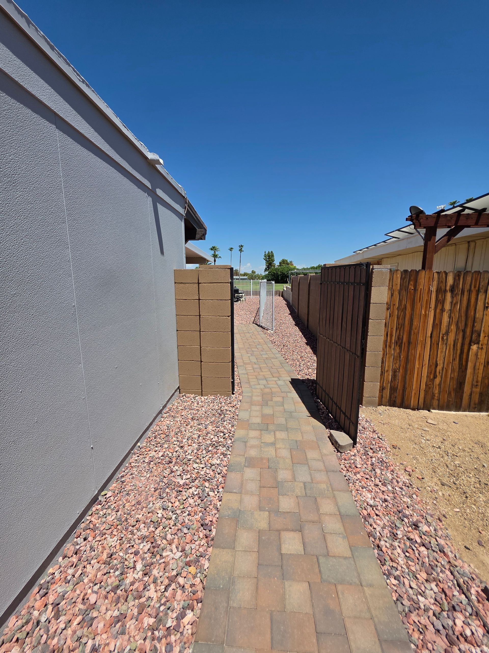 Brick path between two walls, one stucco and the other wood, with red gravel ground cover, under a clear blue sky.