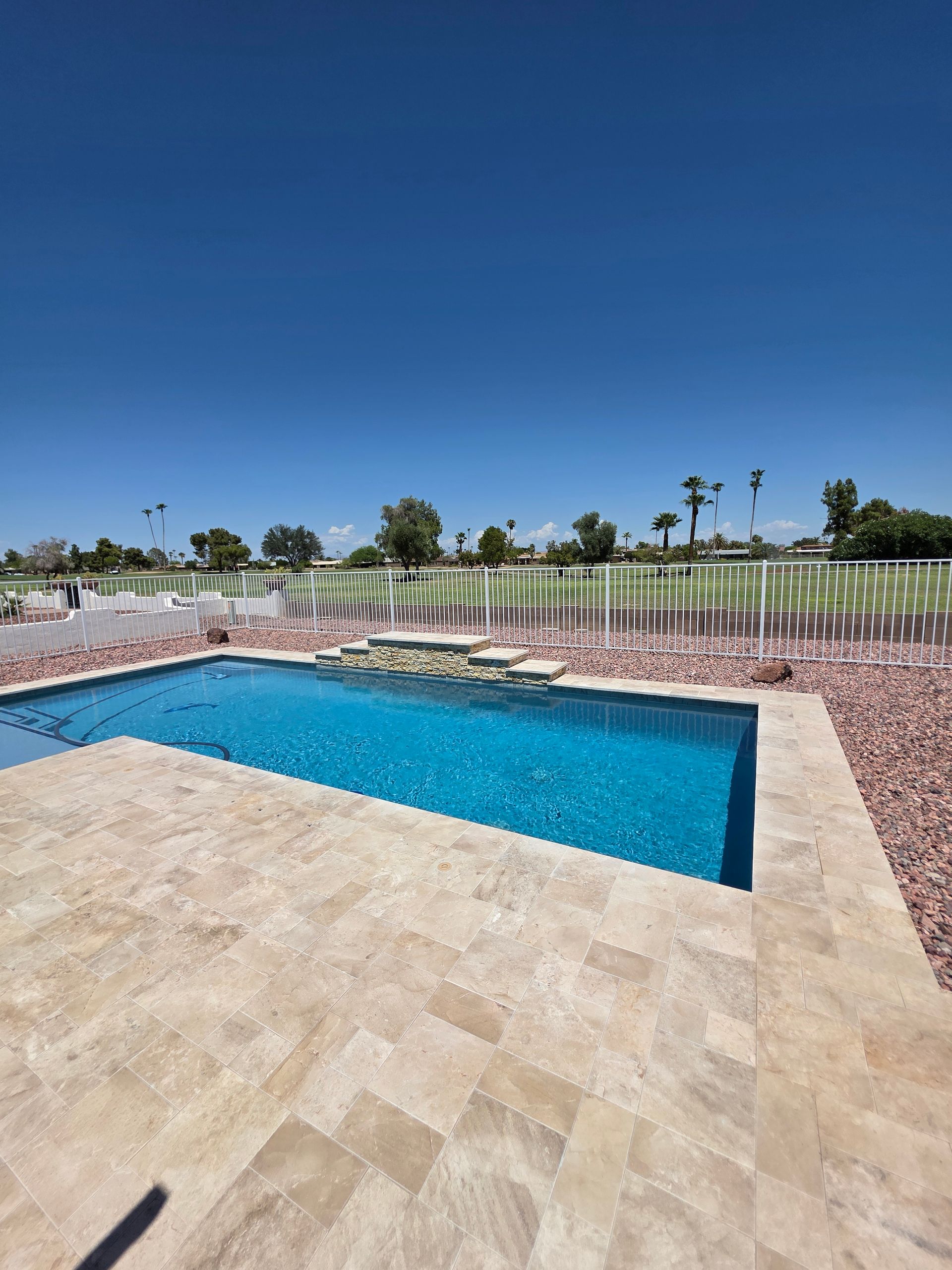 Swimming pool with blue water and beige stone patio under a clear blue sky. Landscaping and trees in the distance.