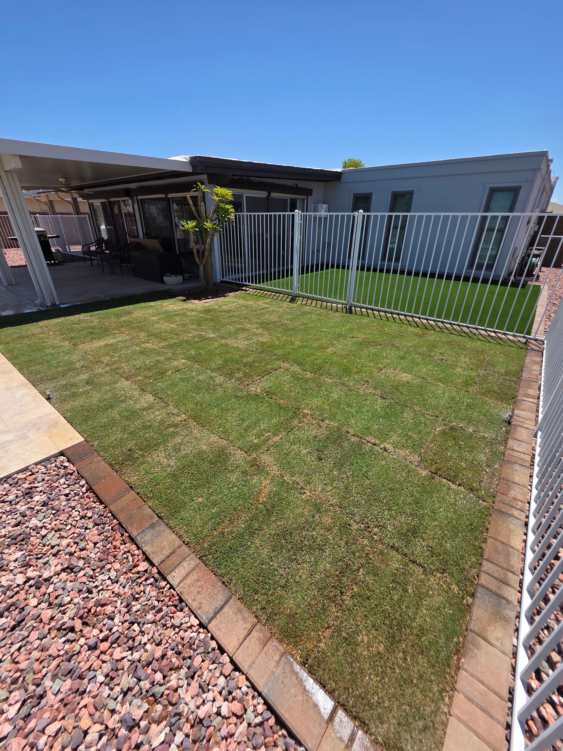 Lush green lawn with brick edging, enclosed by a white fence. A carport provides shade; a building with windows is visible in the background.