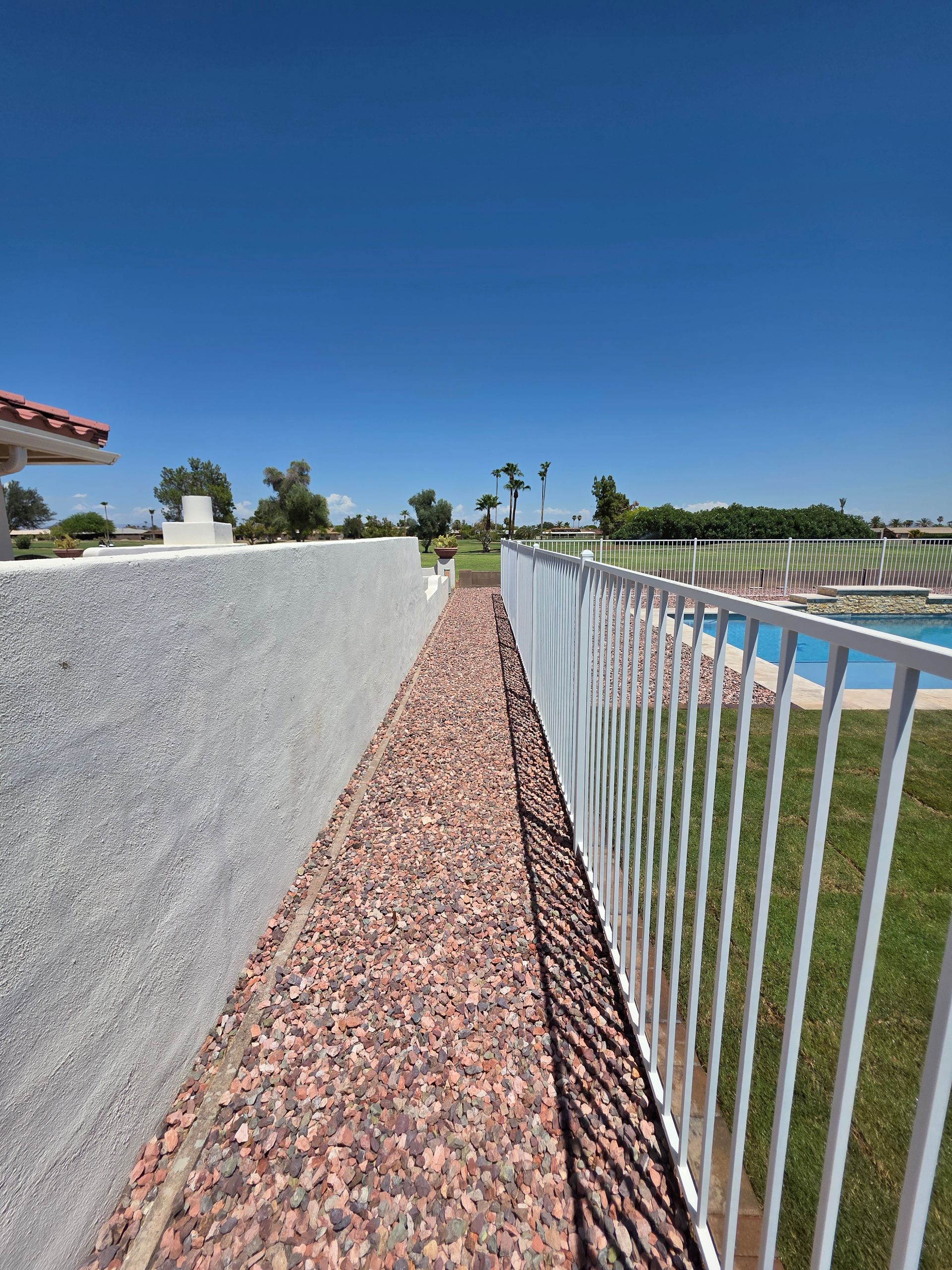 A narrow path with red gravel, bordered by a white fence and a textured white wall, leads to a pool under a bright blue sky.