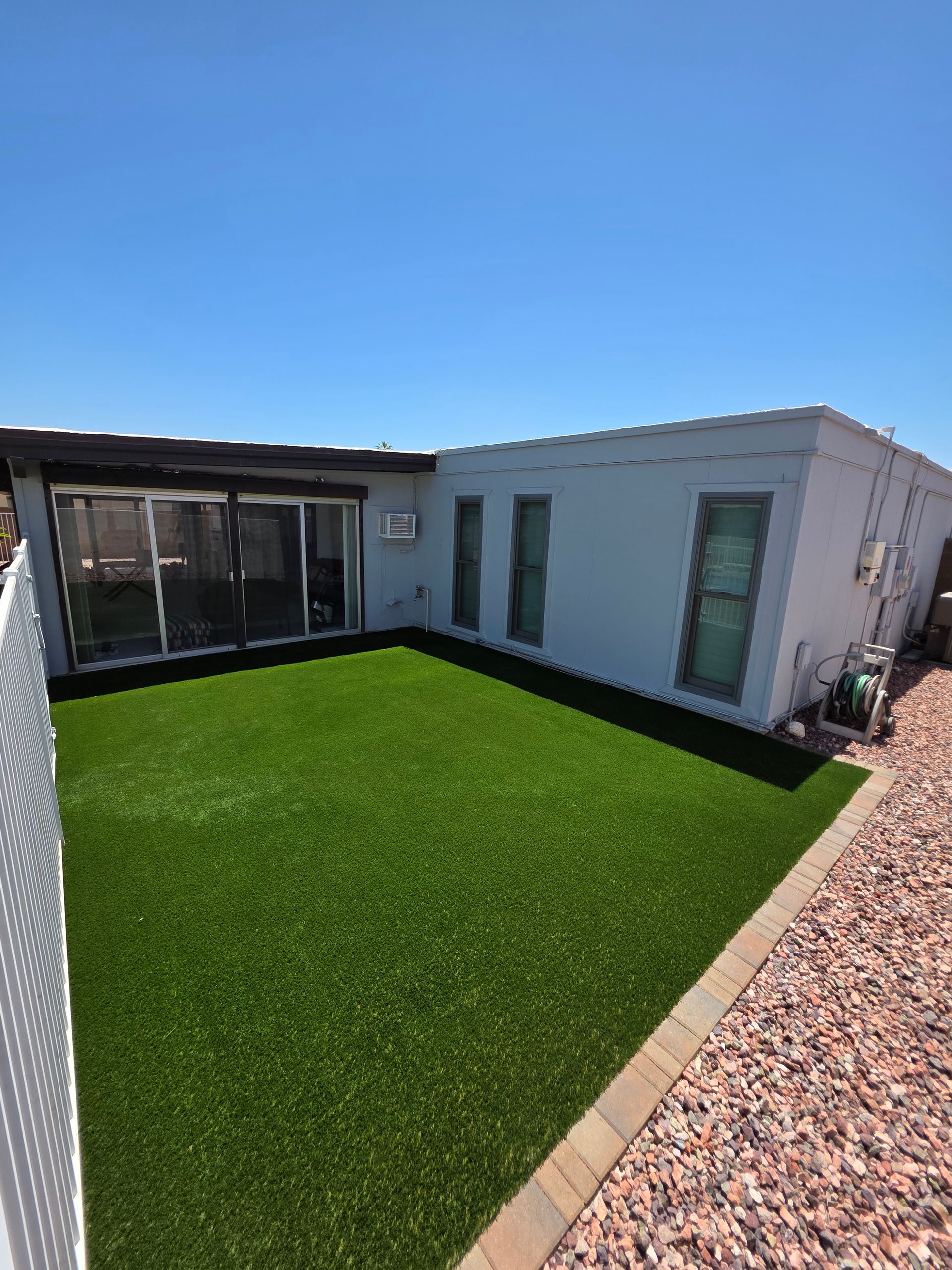 Small backyard with artificial green grass, a light blue house, and a gravel border under a clear blue sky.