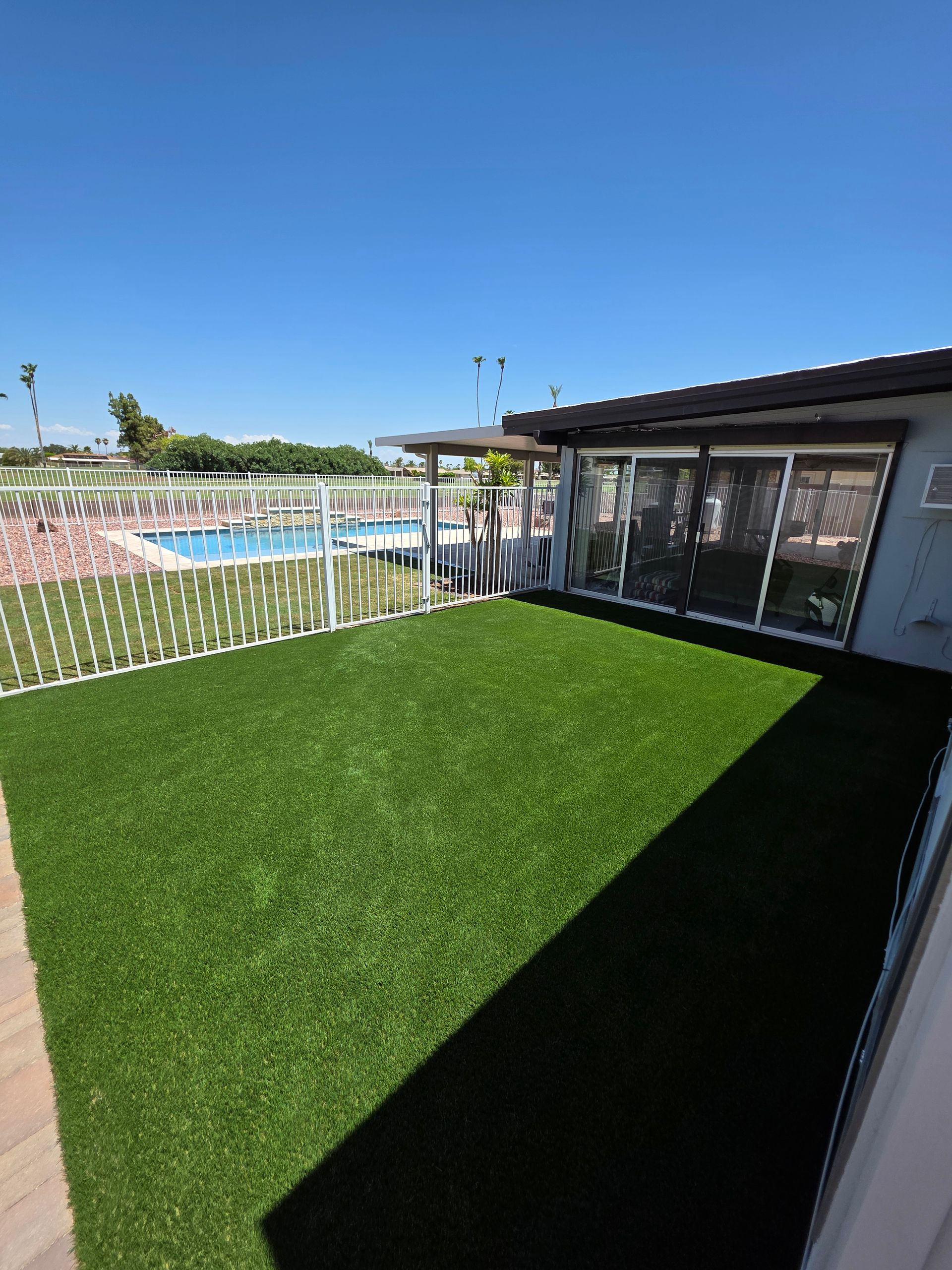 A rooftop patio with green turf, clear blue sky, and a pool area visible beyond a white fence.