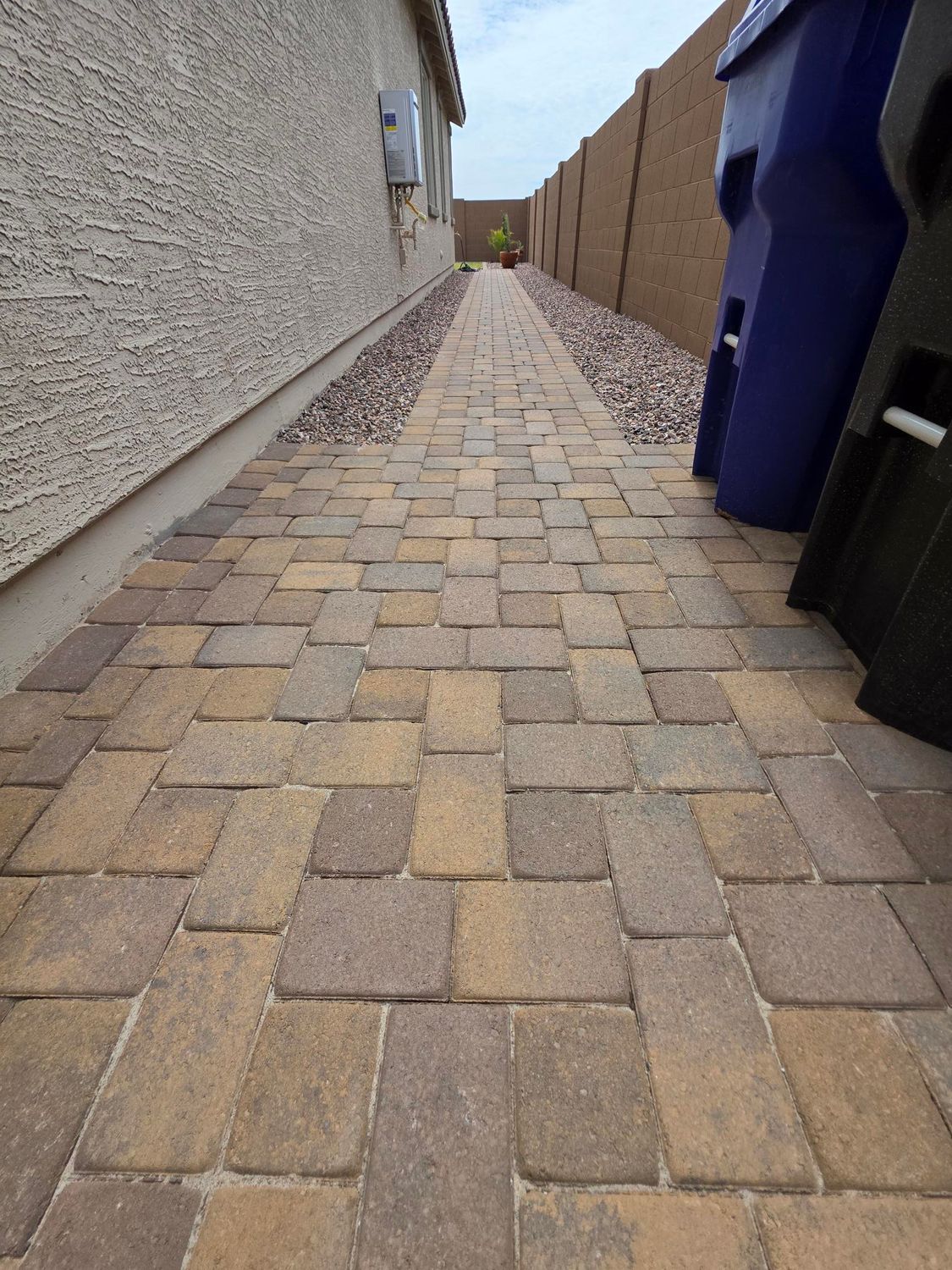 Stone paver pathway between a beige house and a tan wall, with blue trash bins on the right.