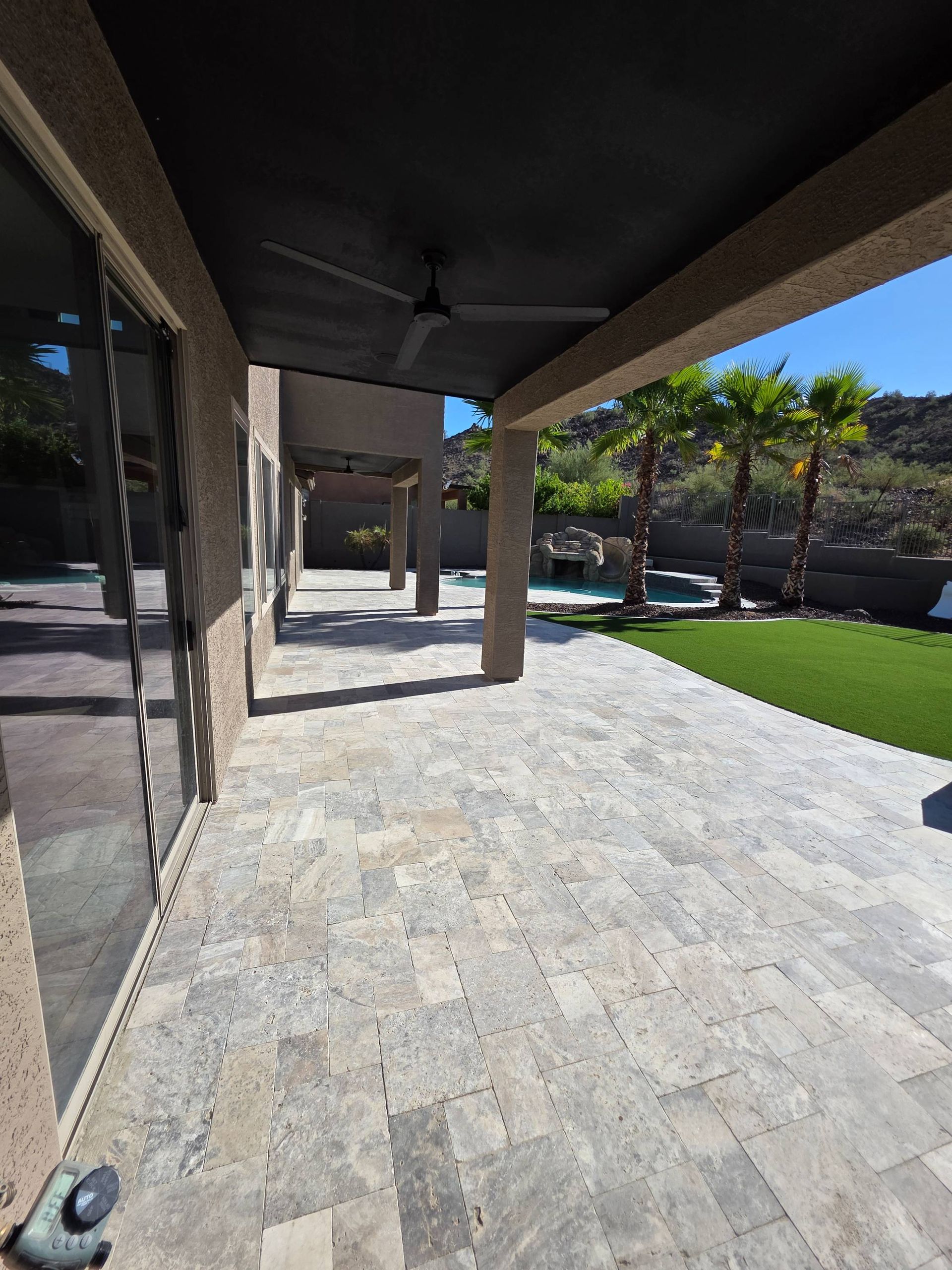 Patio with stone pavers, covered area with ceiling fan, leading to a pool and landscaping.