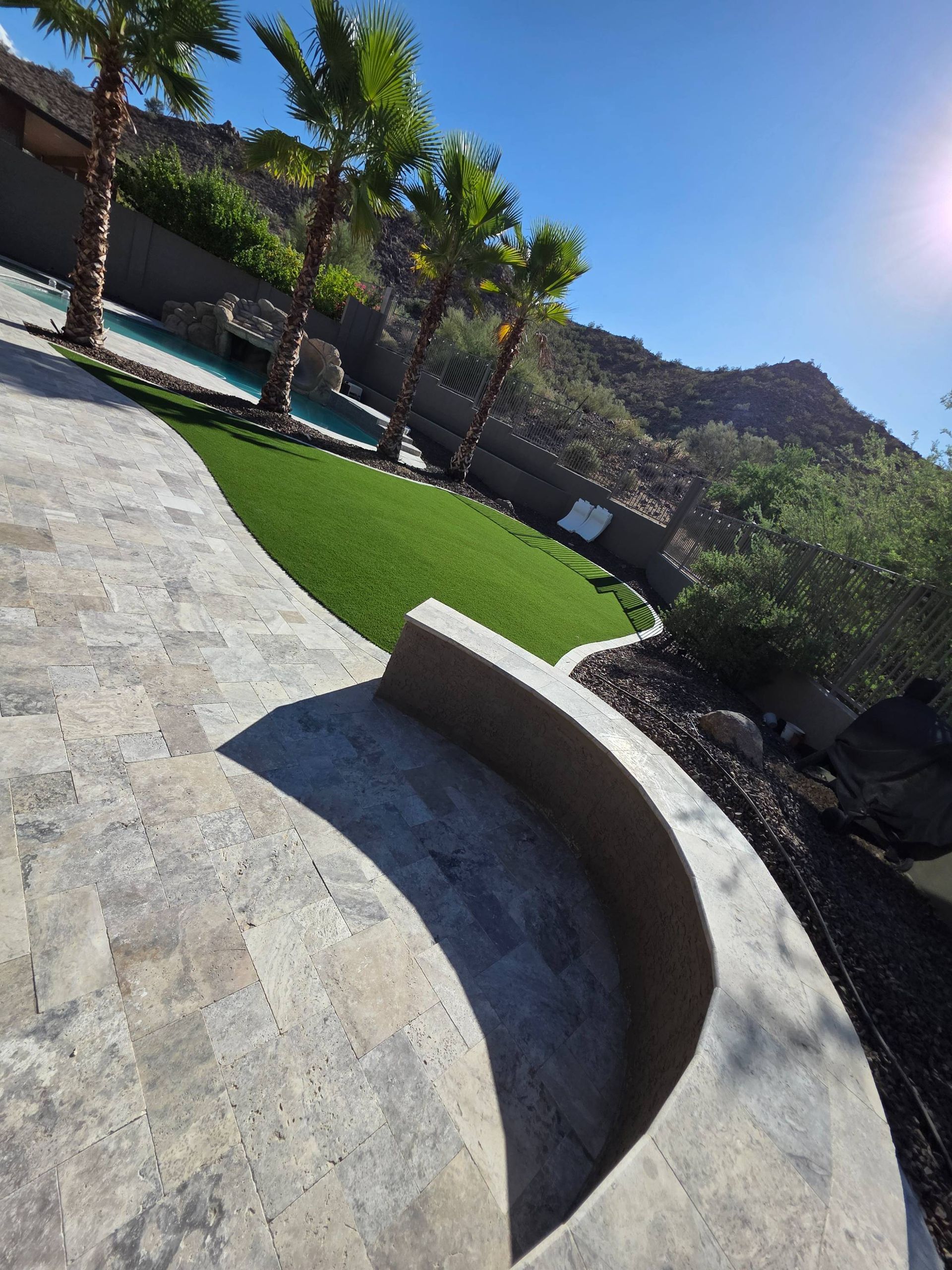 Stone patio with a curved edge overlooking a green lawn, pool, and desert mountain backdrop under a blue sky.