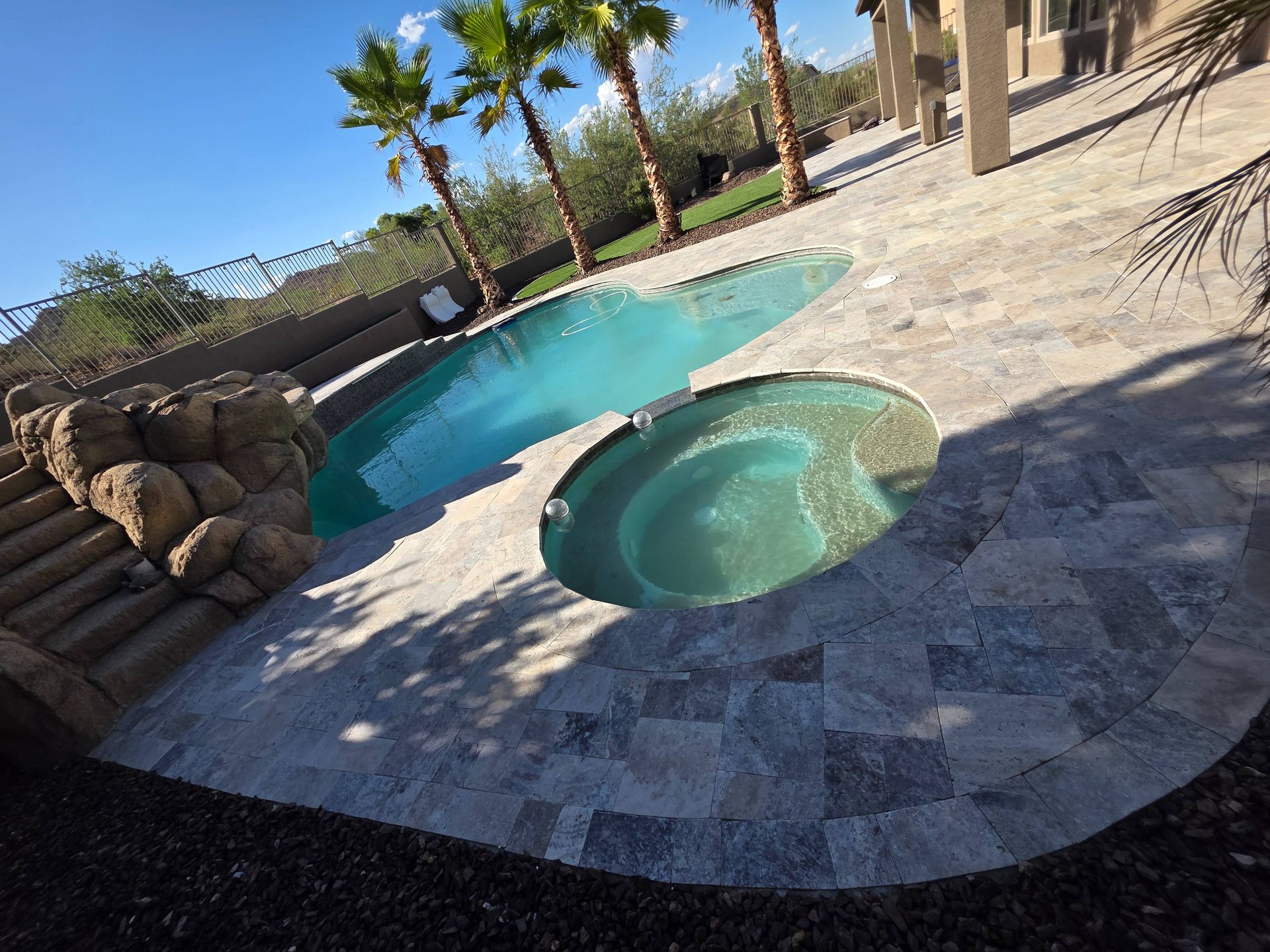 Pool with turquoise water, surrounded by stone patio, palm trees, and sunny sky.