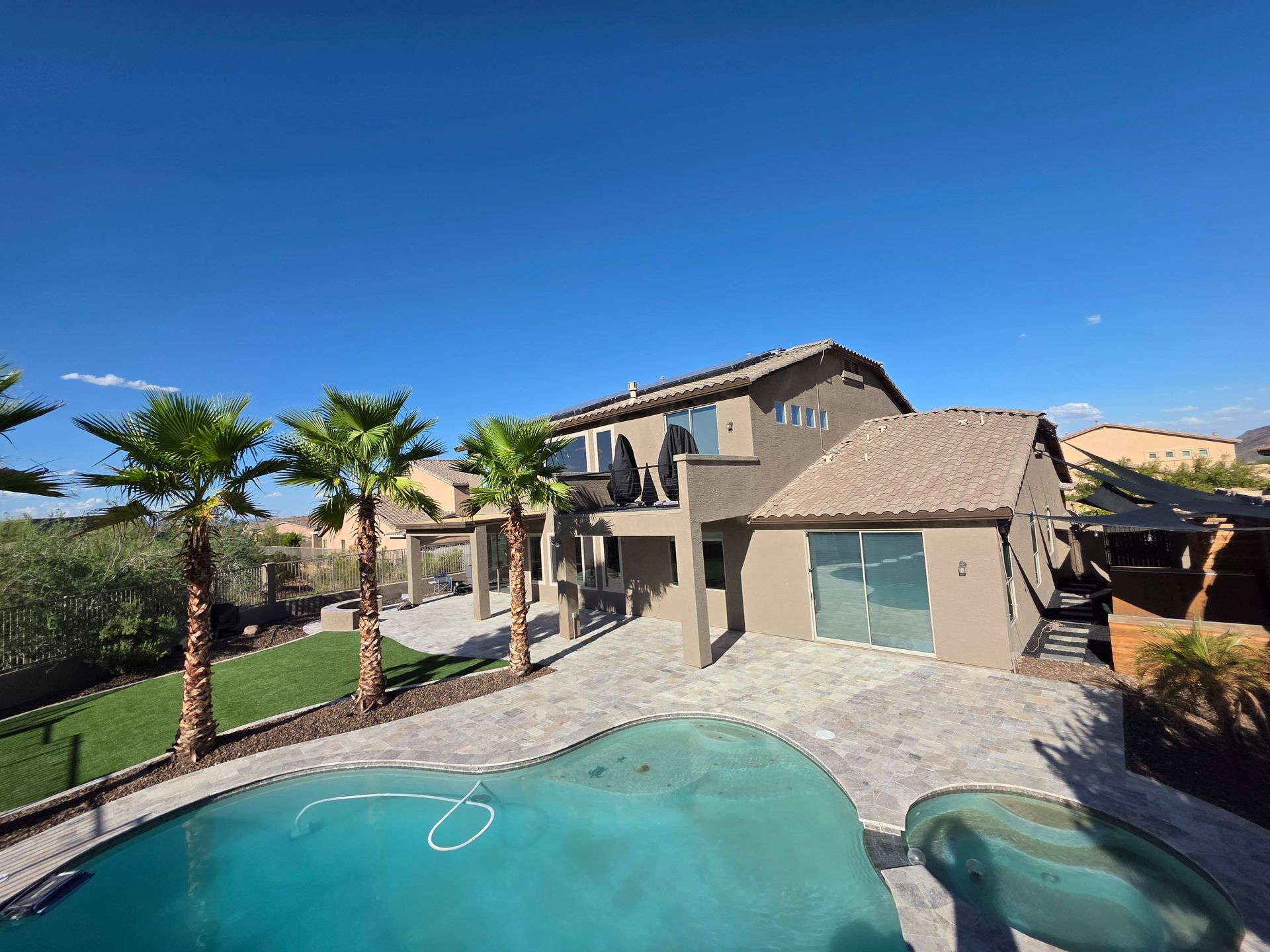 Backyard with pool, patio, and multi-story house under a bright blue sky.