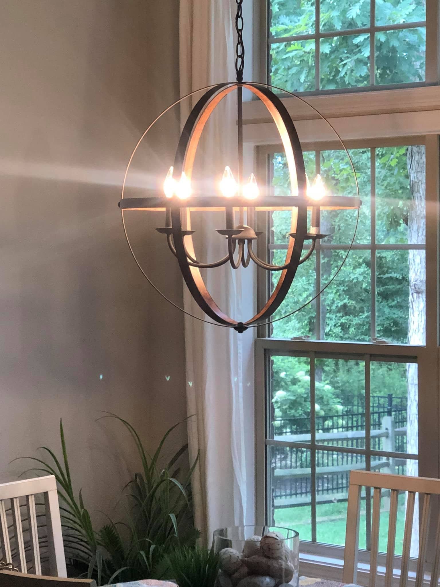 Modern chandelier lit beside a large window in a bright dining room with chairs and plants