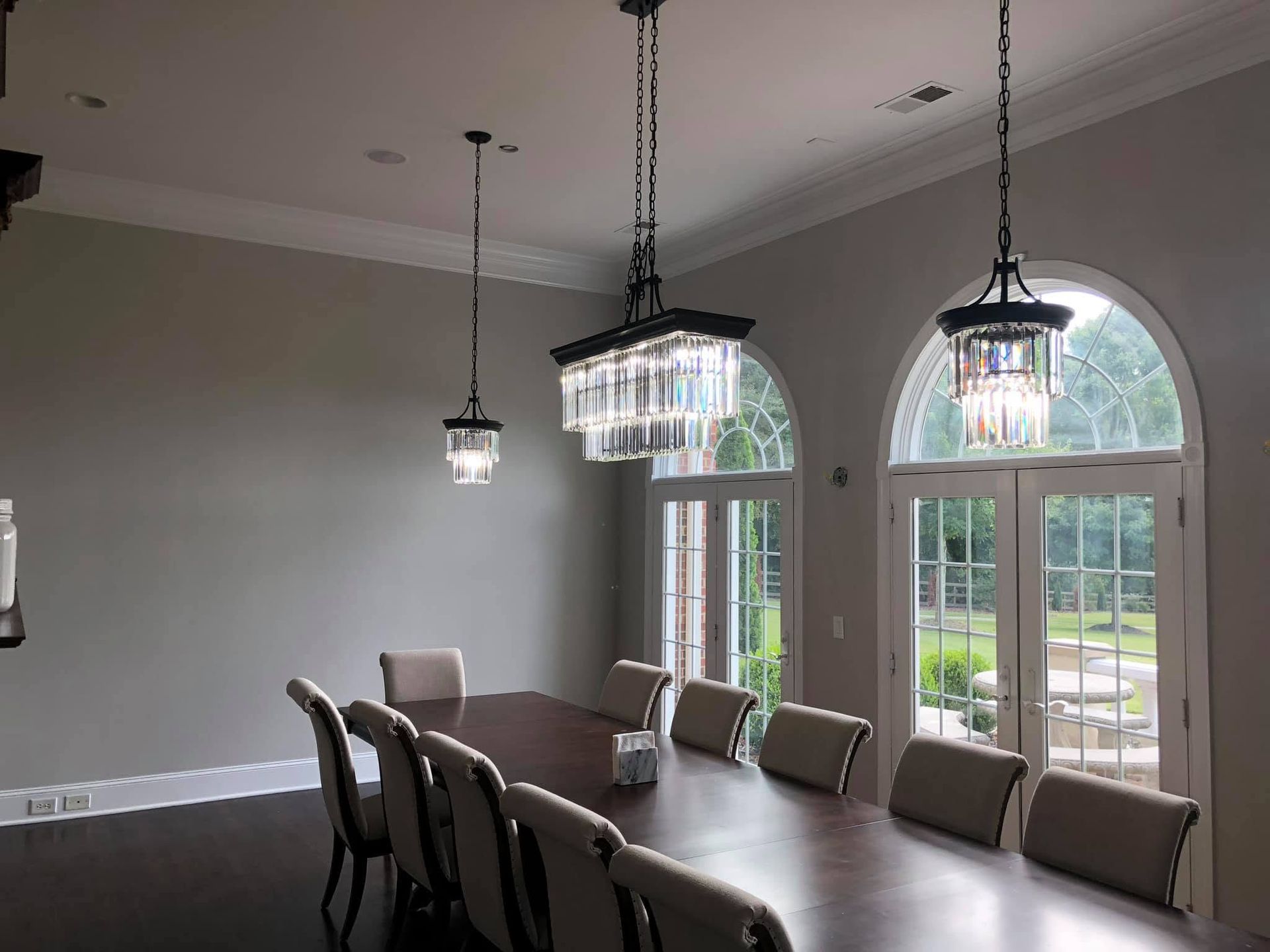 Dining room with long table, upholstered chairs, and pendant lights by arched windows.
