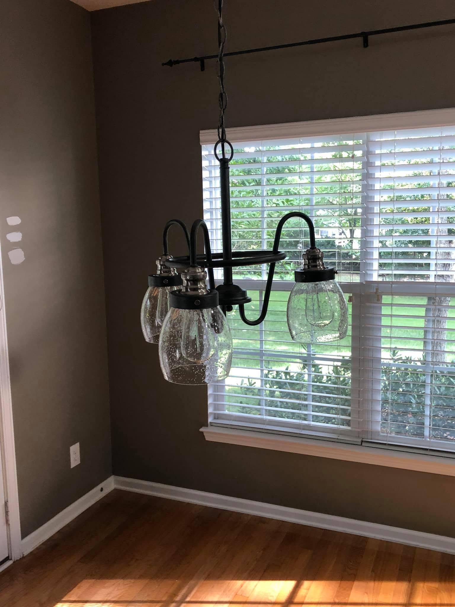 Dining room with a black and glass chandelier hanging by a window with white blinds and hardwood floors.
