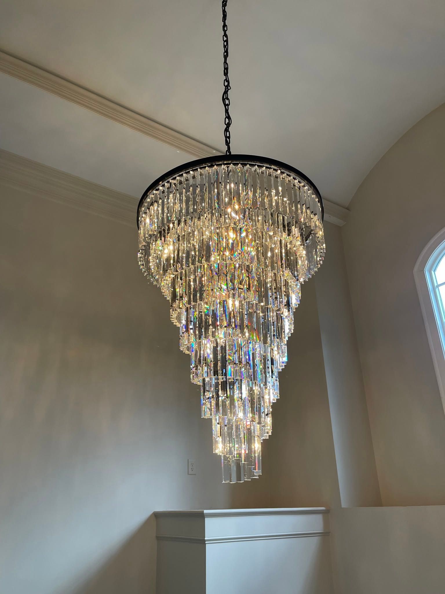 Crystal chandelier hanging in a beige room with a high arched ceiling and window.