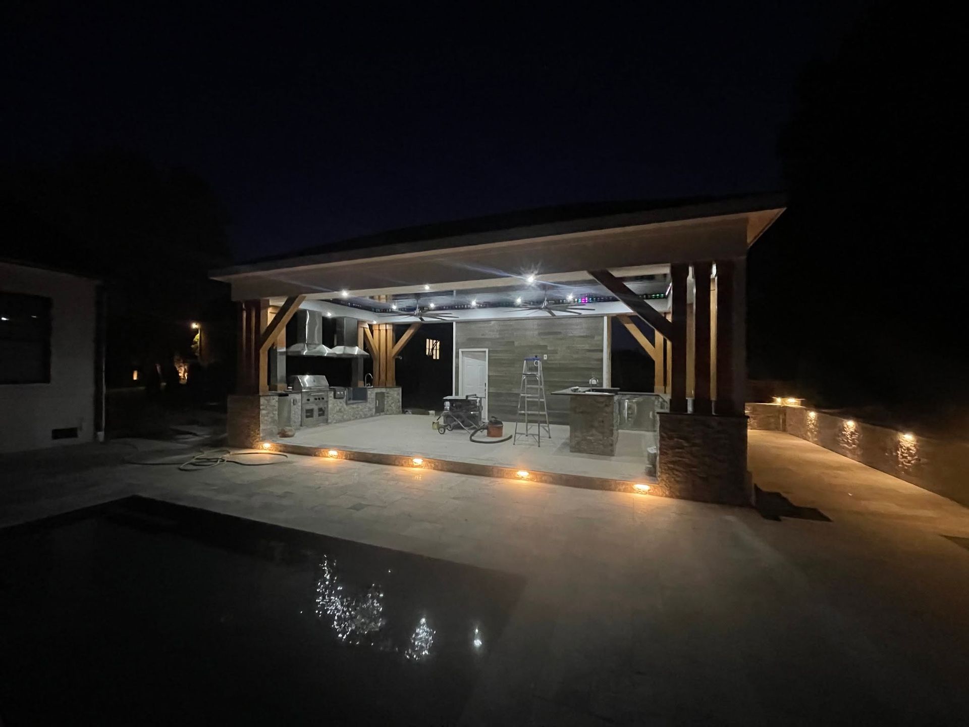 Lit outdoor patio with a roof, bar seating, string lights, and a pool reflecting the lights at night