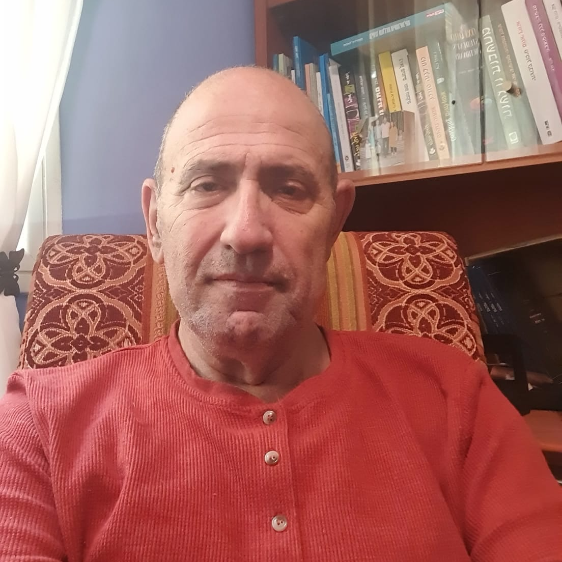 גבר בחולצה אדומה יושב בכיסא, מביט במצלמה. מדף ספרים ברקע.