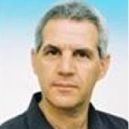 גבר עם שיער אפור קצר, לבוש חולצה כהה, מביט קדימה עם הבעת פנים ניטרלית.