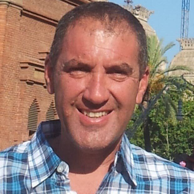 גבר מחייך, לבוש בחולצה כחולה משובצת, מול בניין לבנים ועצי דקל.