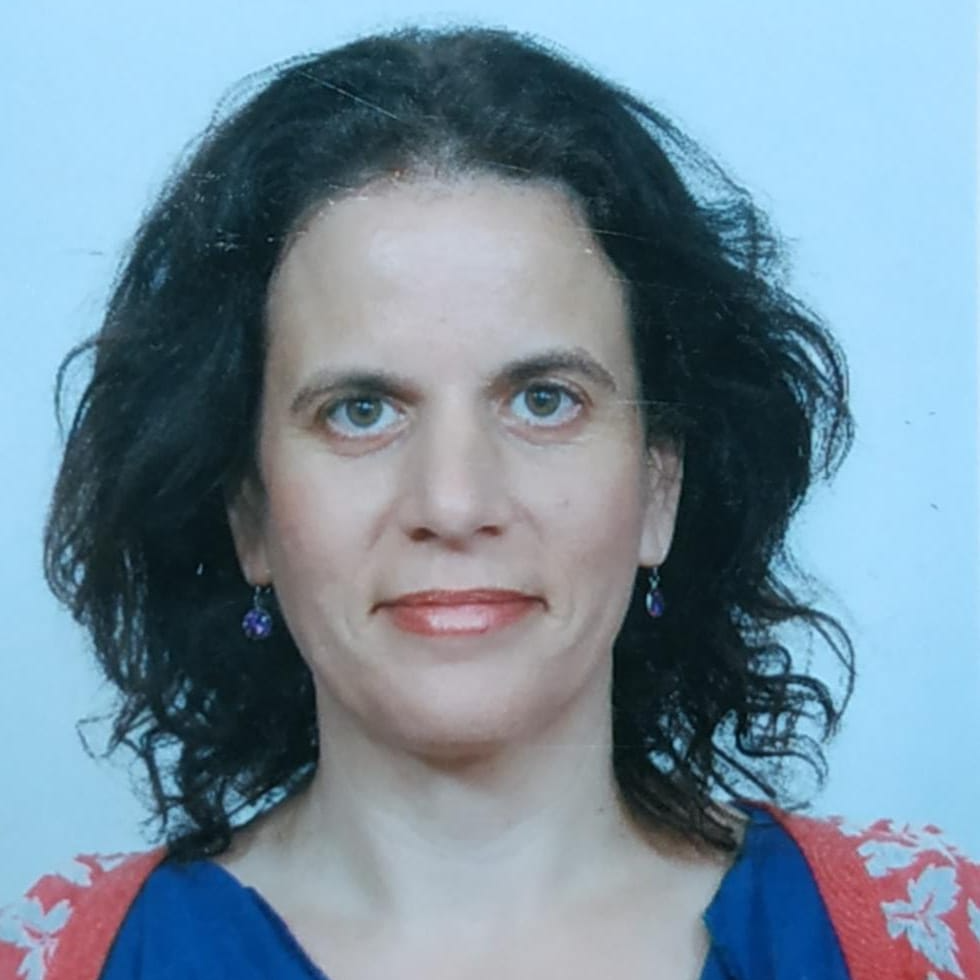אישה עם שיער כהה ועור בהיר מסתכלת למצלמה, לובשת חולצה כחולה וסוודר אדום, רקע פשוט.