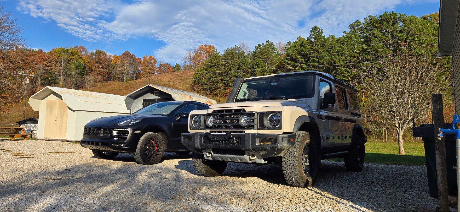 Two cars parked on gravel; one black and one beige/black with a shed and trees in the background.