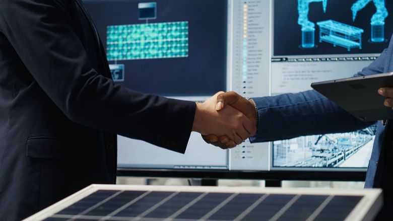 Two people shake hands in front of computer screens, suggesting a business agreement.