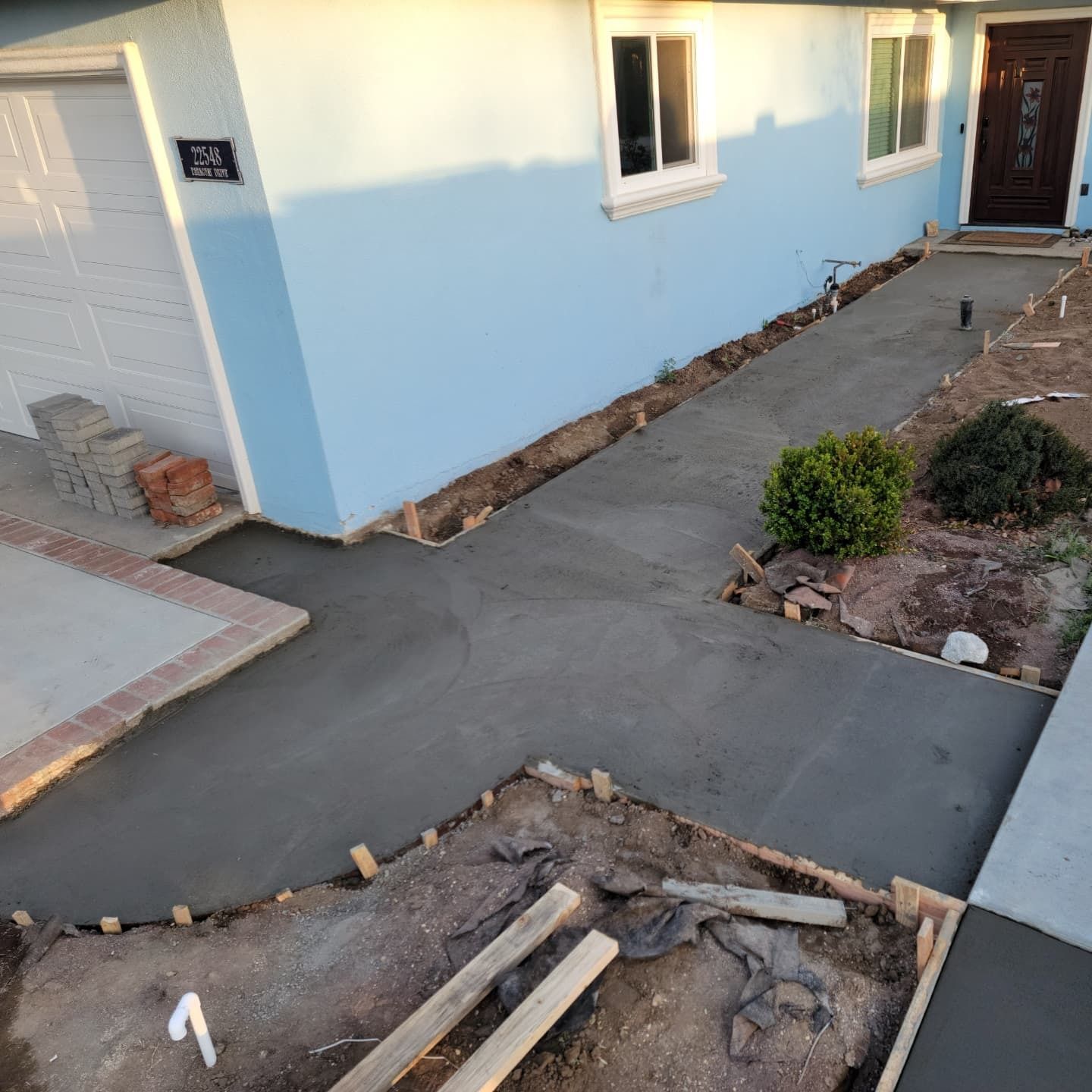 Freshly poured concrete pathway in front of a blue house, with wooden framing and small bushes.