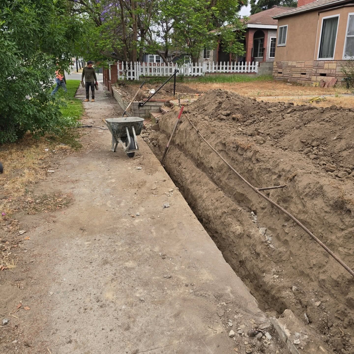 A long trench dug in the dirt beside a sidewalk; a person and a wheelbarrow are nearby.