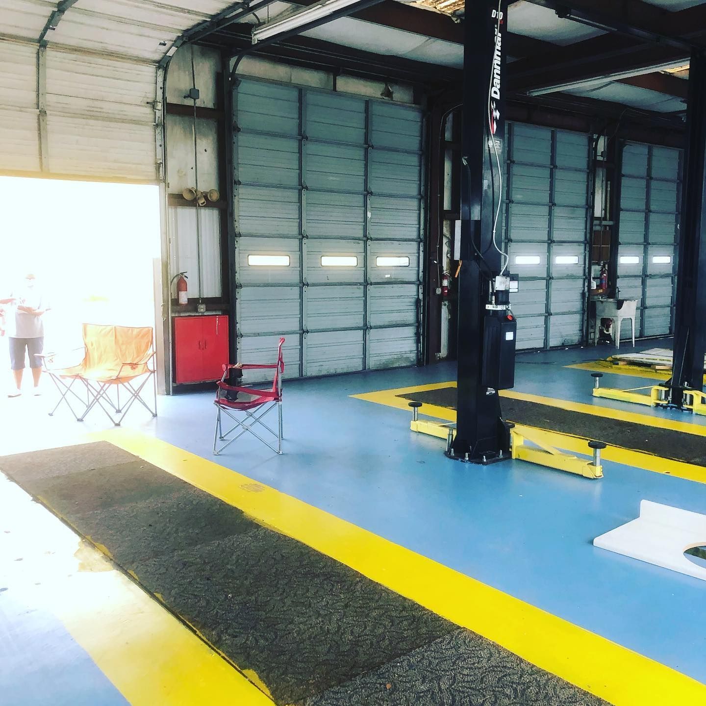 Interior of a garage with blue floors, yellow lift arms, a black mat, and folding chairs near an open bay door