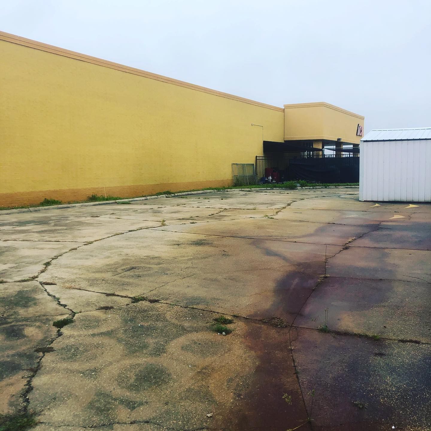 Empty cracked concrete lot beside a long yellow wall and a white shed under an overcast sky