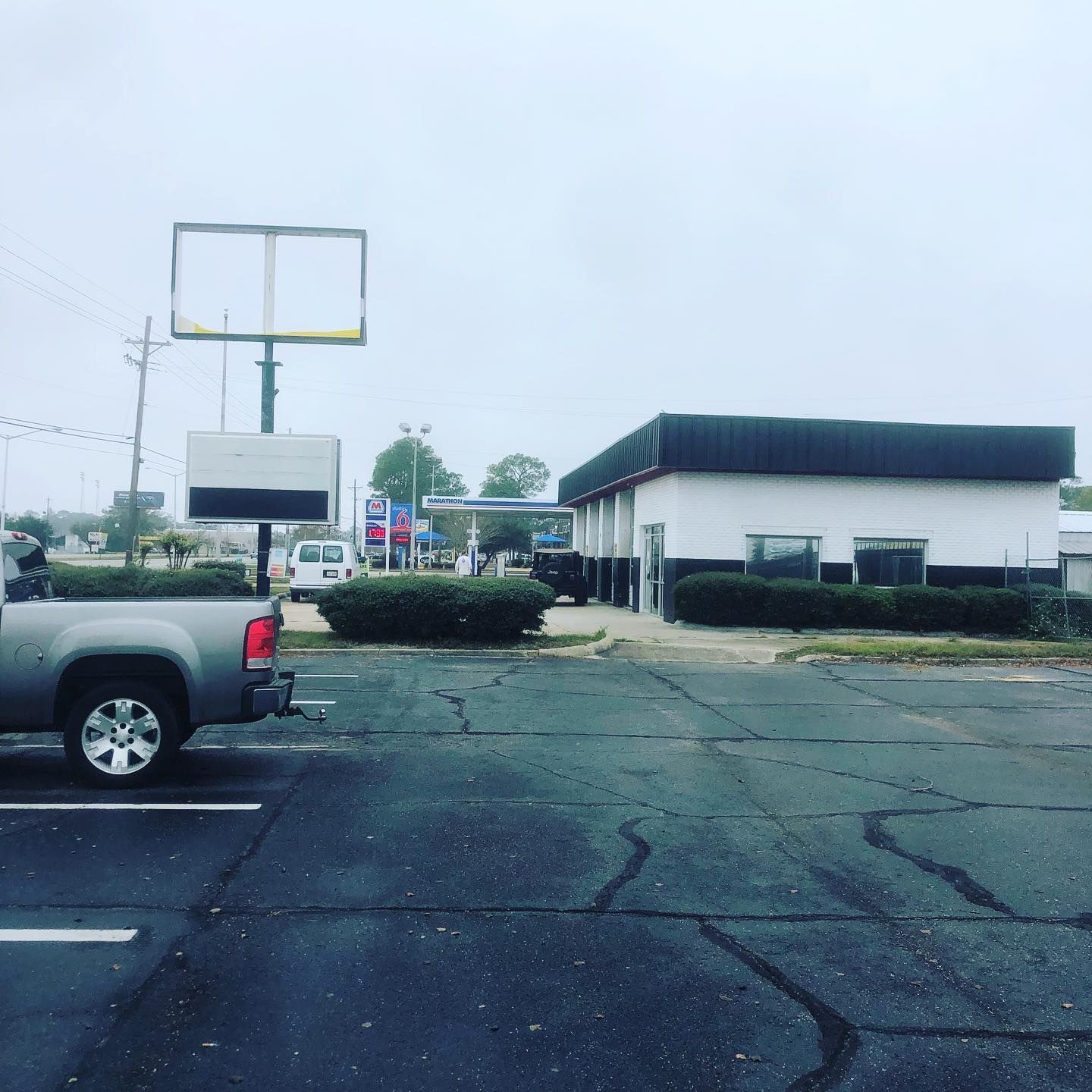 Empty parking lot with a gray pickup truck, cracked asphalt, and a low commercial building under a cloudy sky