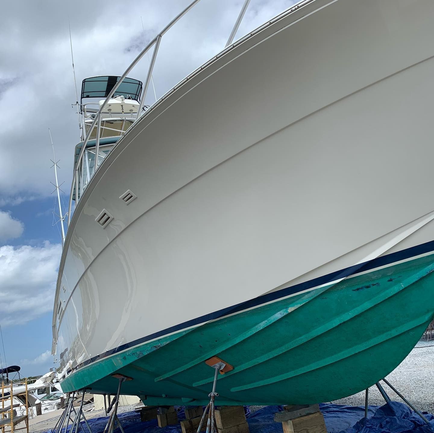 White yacht hull with turquoise underside on a boat stand at a marina under a cloudy sky