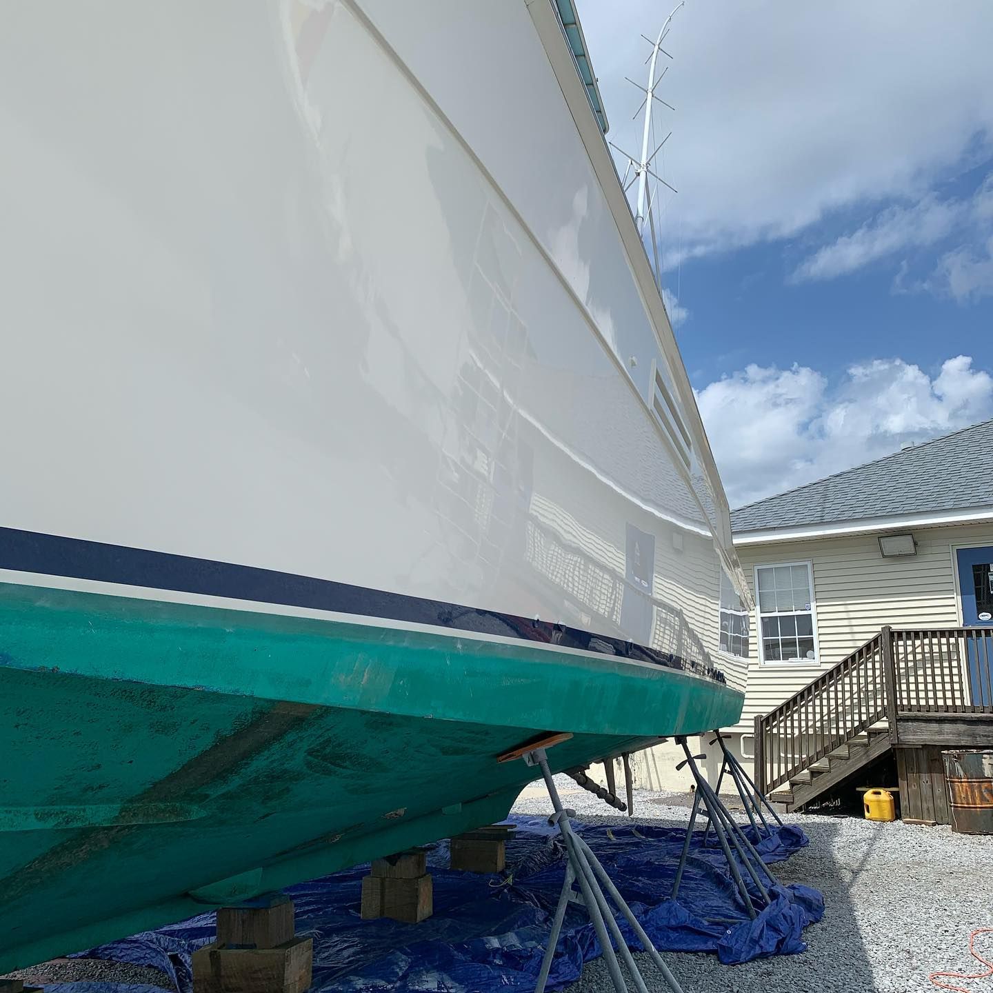 White boat hull on supports with teal lower hull beside a house and stairs on a gravel lot