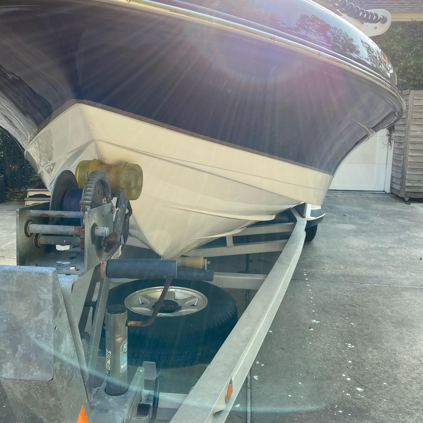 Boat hull on a trailer in a driveway, with a bright sun flare overhead.