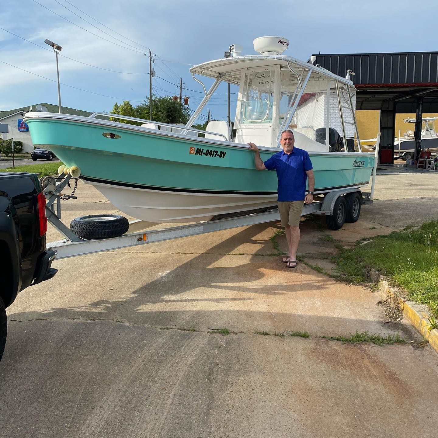 Man standing beside a teal boat on a trailer in a driveway near a waterfront building.