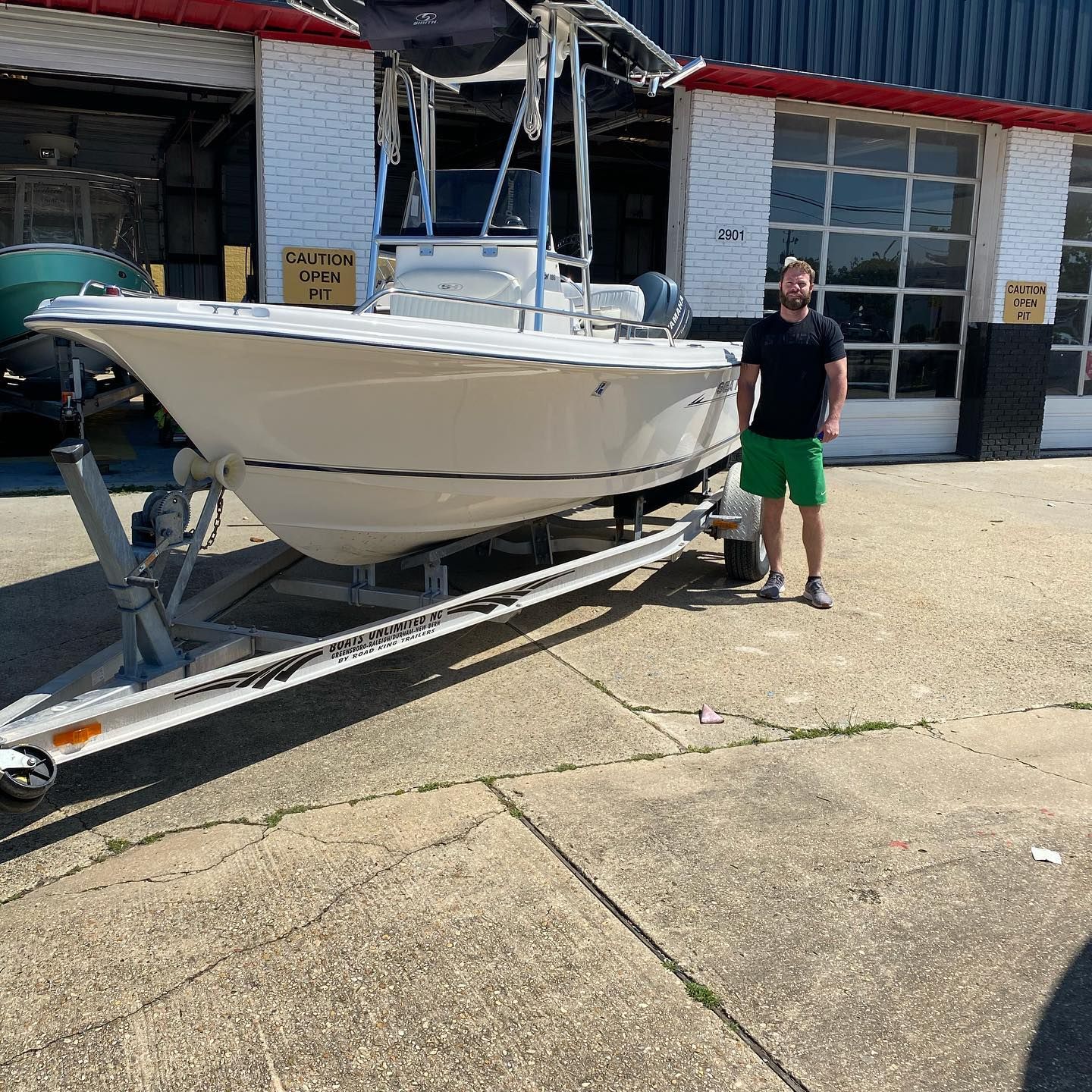 Man standing beside a white boat on a trailer outside a boat shop on a sunny day