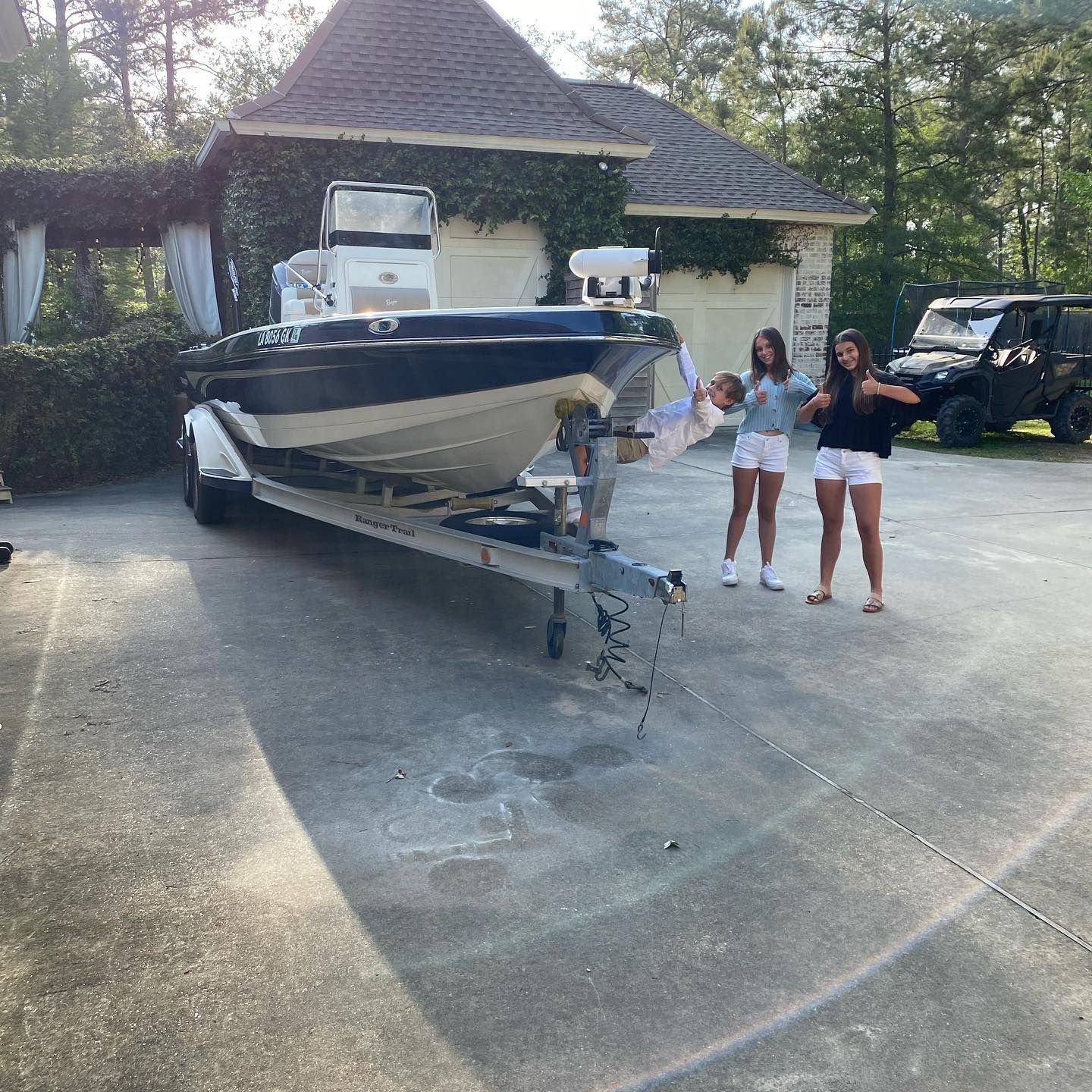 Boat on trailer in a driveway, with two people standing beside it near a house and parked vehicles.