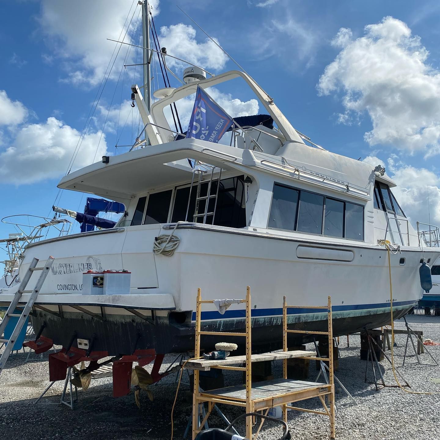 White yacht on stands in a gravel lot under a blue sky with clouds, seen from the stern side.